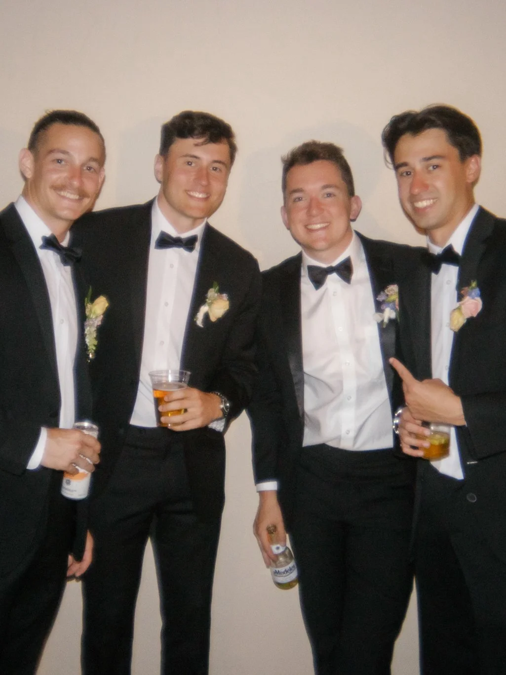 Four men in tuxedos with bow ties smiling and holding drinks, standing against a beige wall.