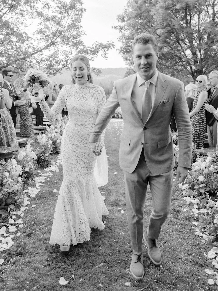 A newlywed couple walking hand-in-hand down an outdoor aisle, holding a bouquet, with guests clapping and celebrating around them, trees in the background.