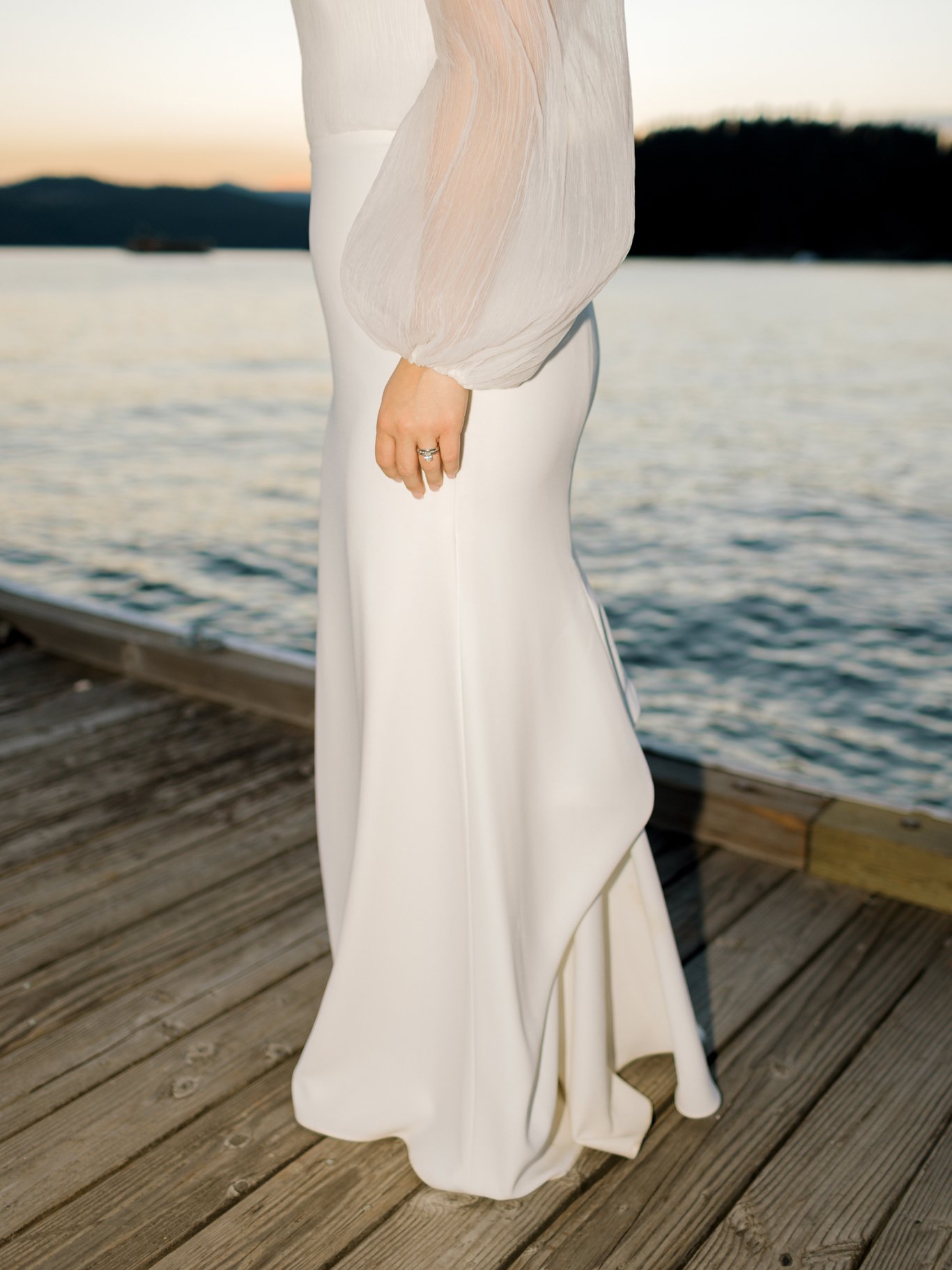 A person standing on a wooden dock near water at sunset, wearing a long, flowing white dress and a sheer, long-sleeved blouse, with their left hand resting at their side, showing a ring on their finger at the Hagadone Event Center in Coeur d'Alene.