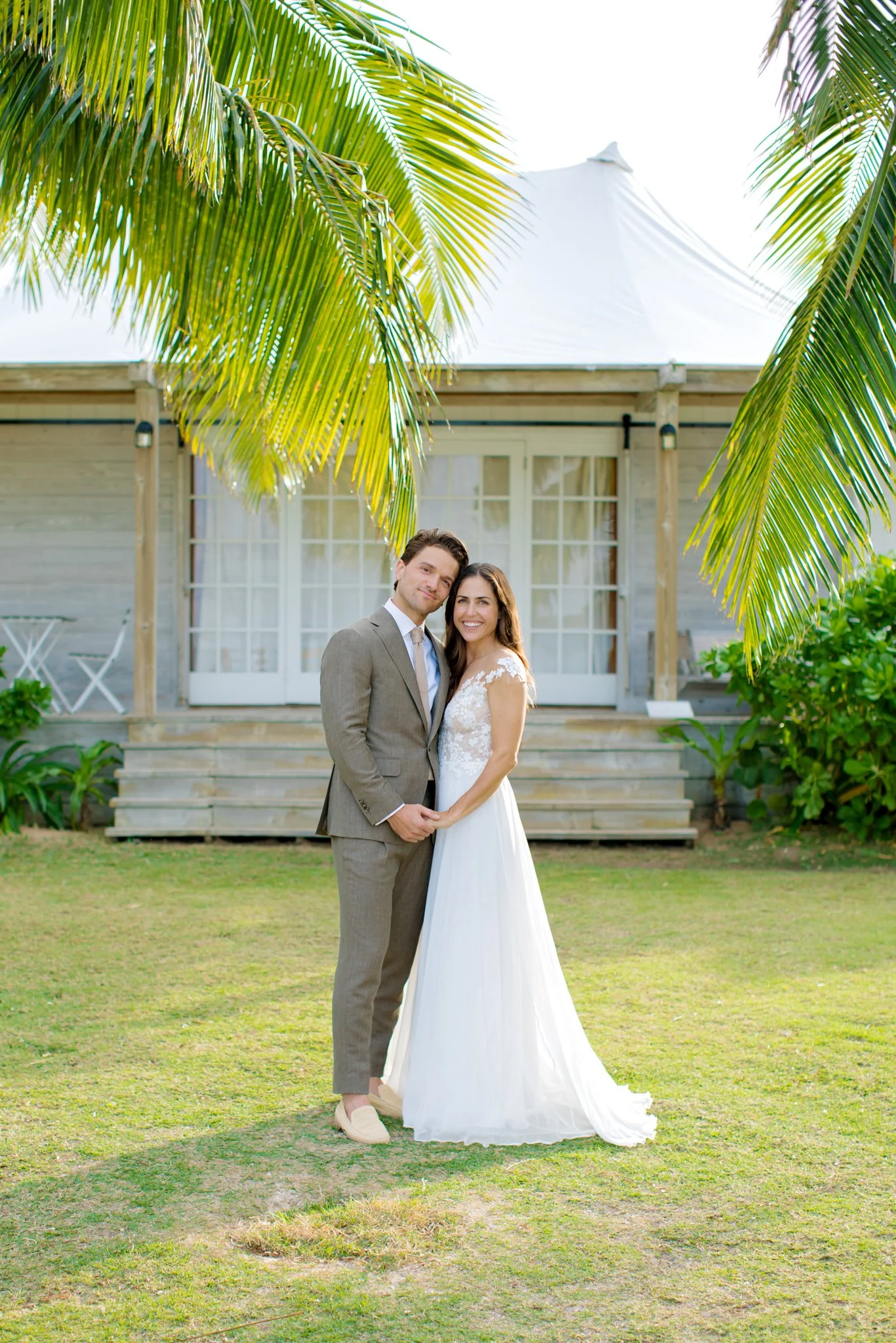 A newlywed couple standing on a lawn in front of a wooden house with a white roof, holding hands and smiling. Palm trees frame the scene at The Other Side Hotel Eleuthera during a luxury Bahamas destination wedding.