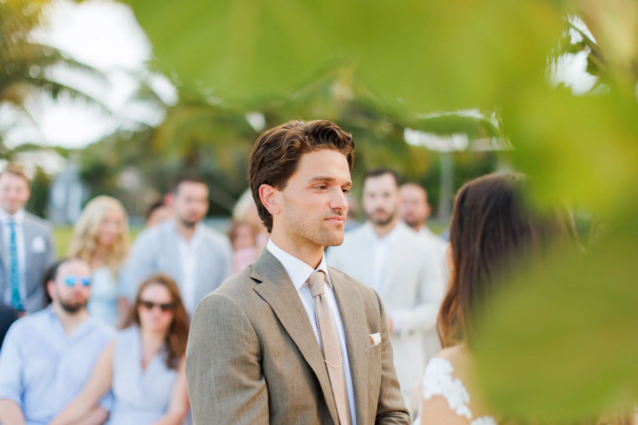 A man in a gray suit and tie gazes at a woman during a wedding ceremony outdoors, with guests in the background and greenery around.