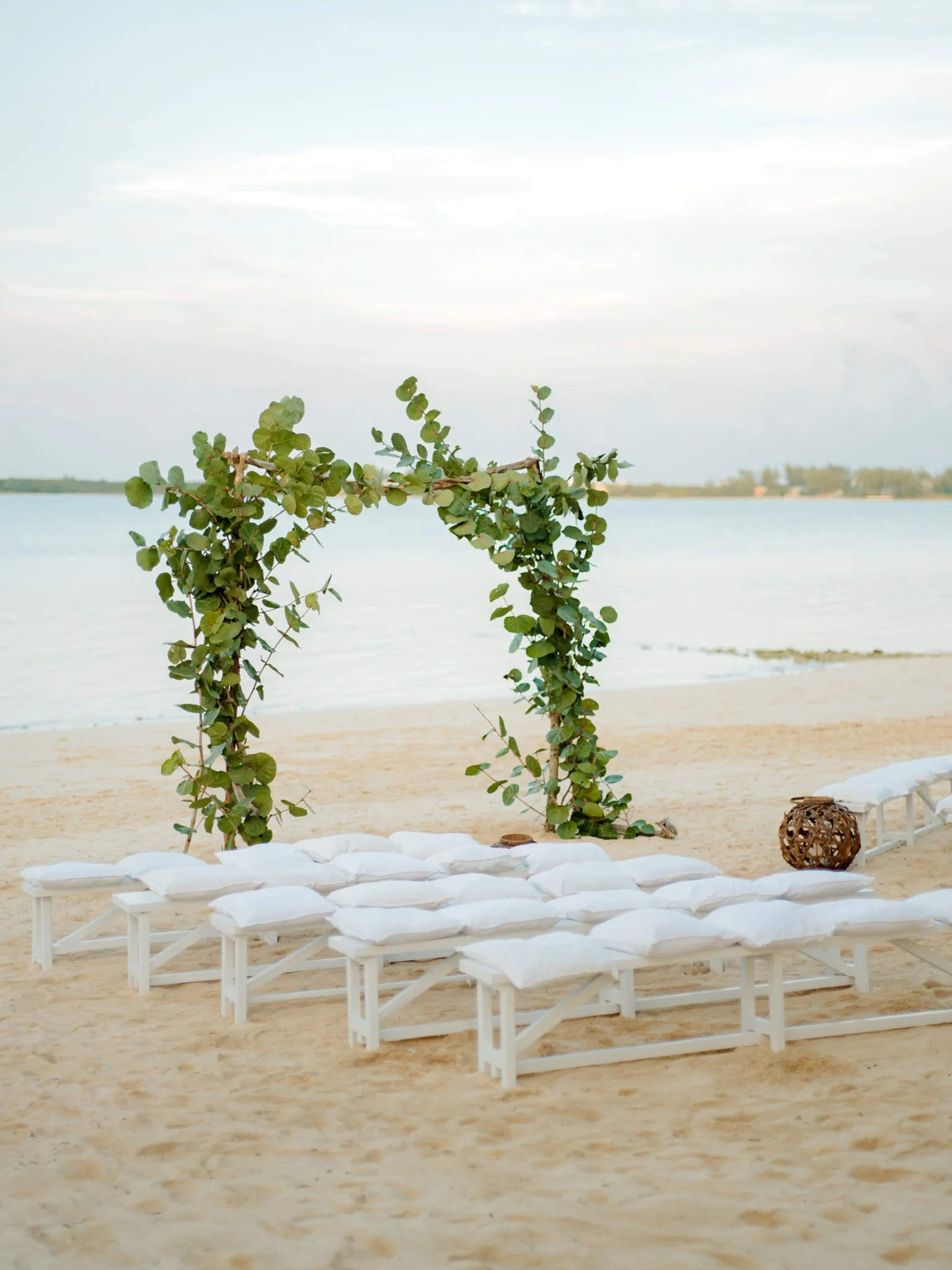 Beach wedding setup with white chairs and a leafy arch over the water.