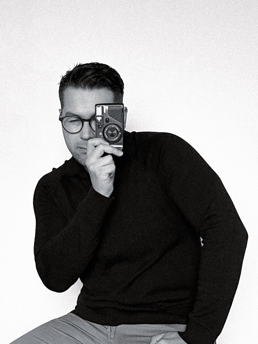 A person with short dark hair and glasses holding a camera, taking a mirror selfie against a plain light background.