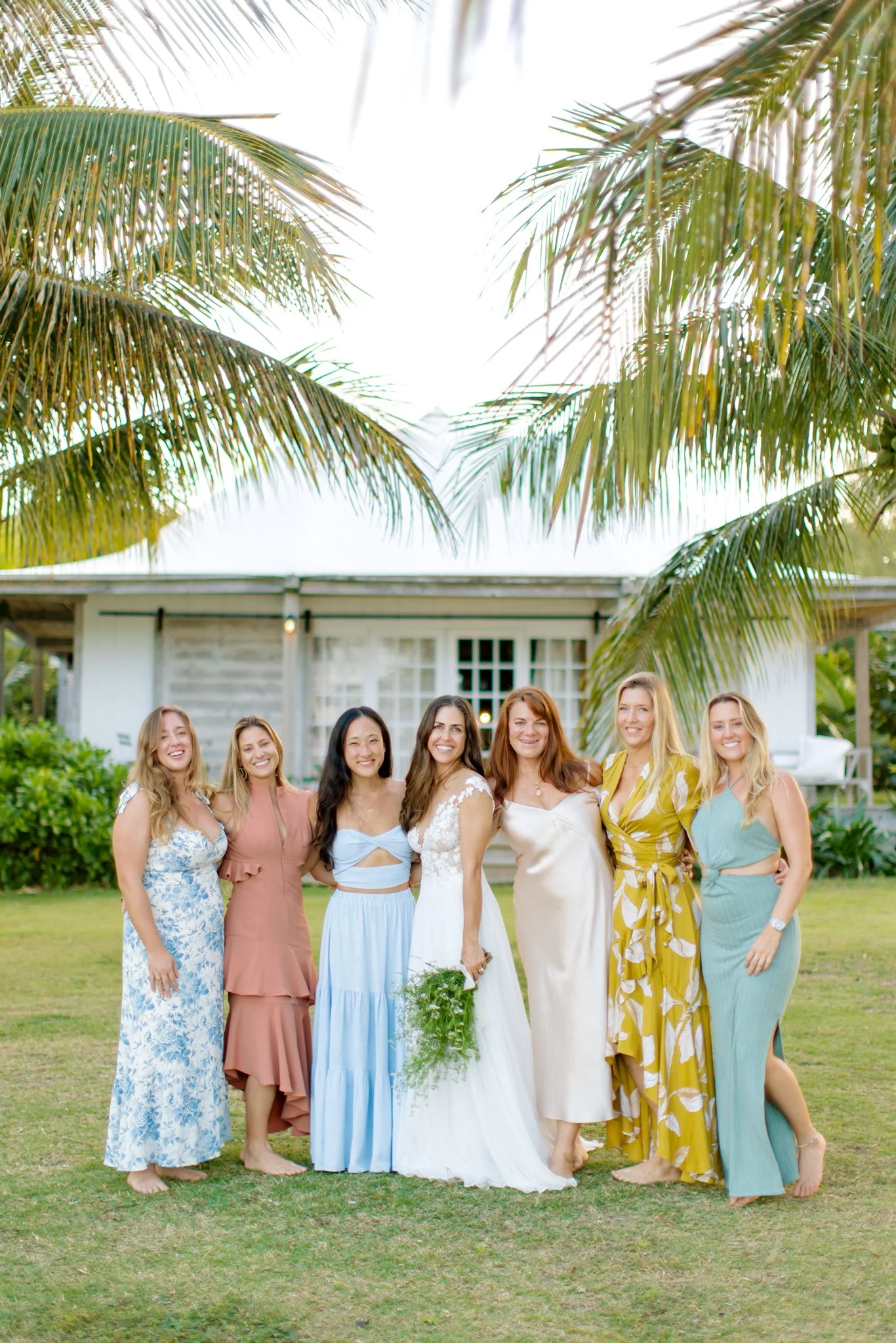 Group of women in colorful dresses standing on grass in front of a white house with palm trees overhead at The Other Side Hotel Eleuthera during a luxury Bahamas destination wedding.