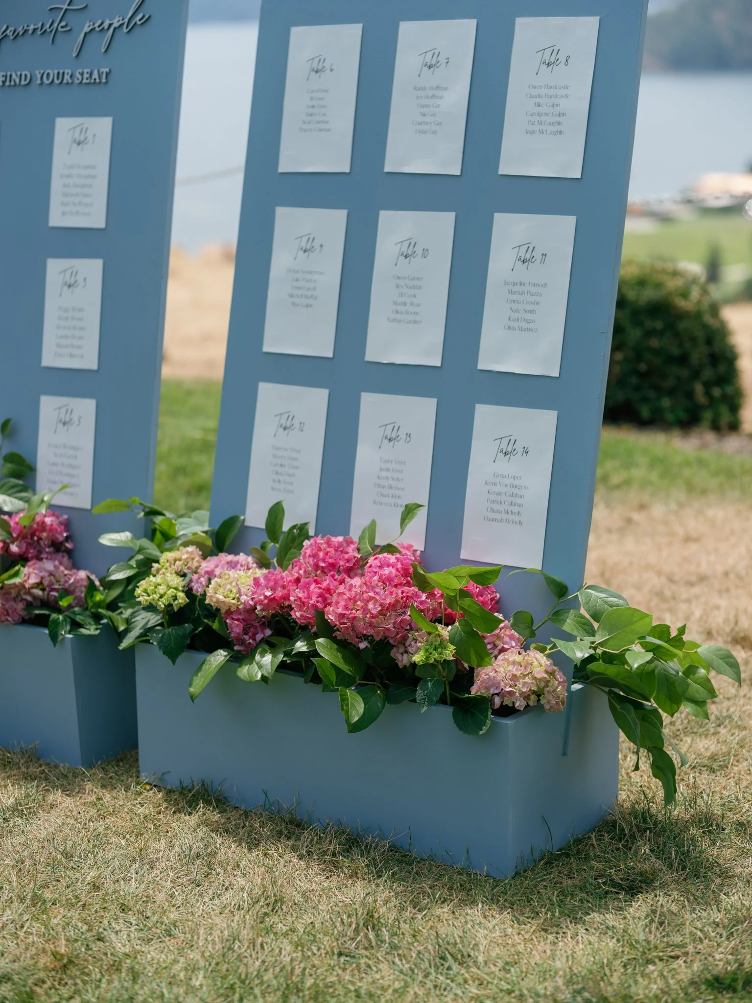 A seating chart for an event displayed on a light blue board, with handwritten table numbers and guest names, placed outdoors on grass with pink and white flowers in blue planters at the base at Elk Point wedding above Lake Coeur d’Alene.