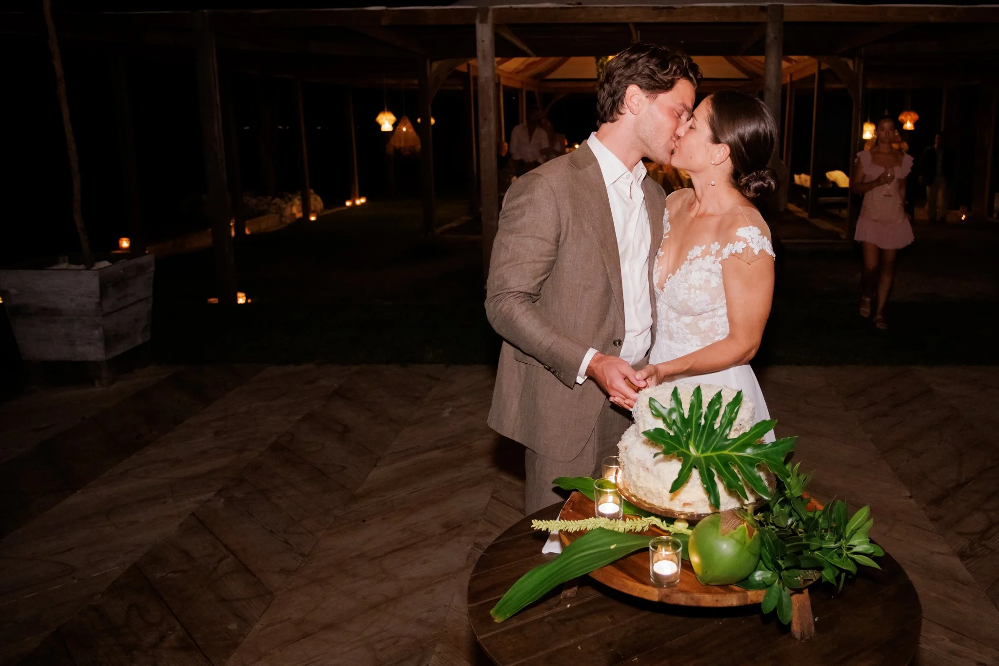 A couple in wedding attire kissing at their wedding reception, with a wedding cake and candles on a table in front of them.