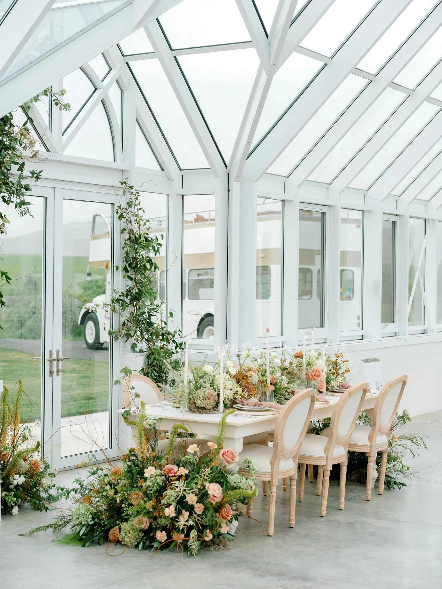 A bright, modern greenhouse with a white table and elegant chairs set for a meal, decorated with floral centerpieces and surrounded by lush plants and flowers.