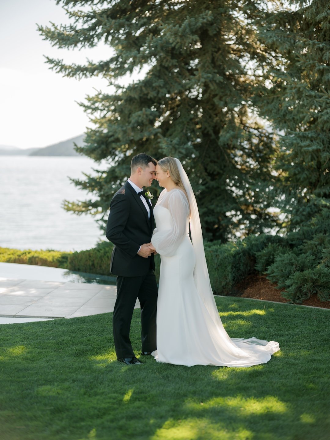 A bride and groom are standing close together outdoors, with their foreheads touching, during a wedding photoshoot. The groom is wearing a black tuxedo with a bow tie, and the bride is in a long white wedding gown with a veil. They are on a grassy ar