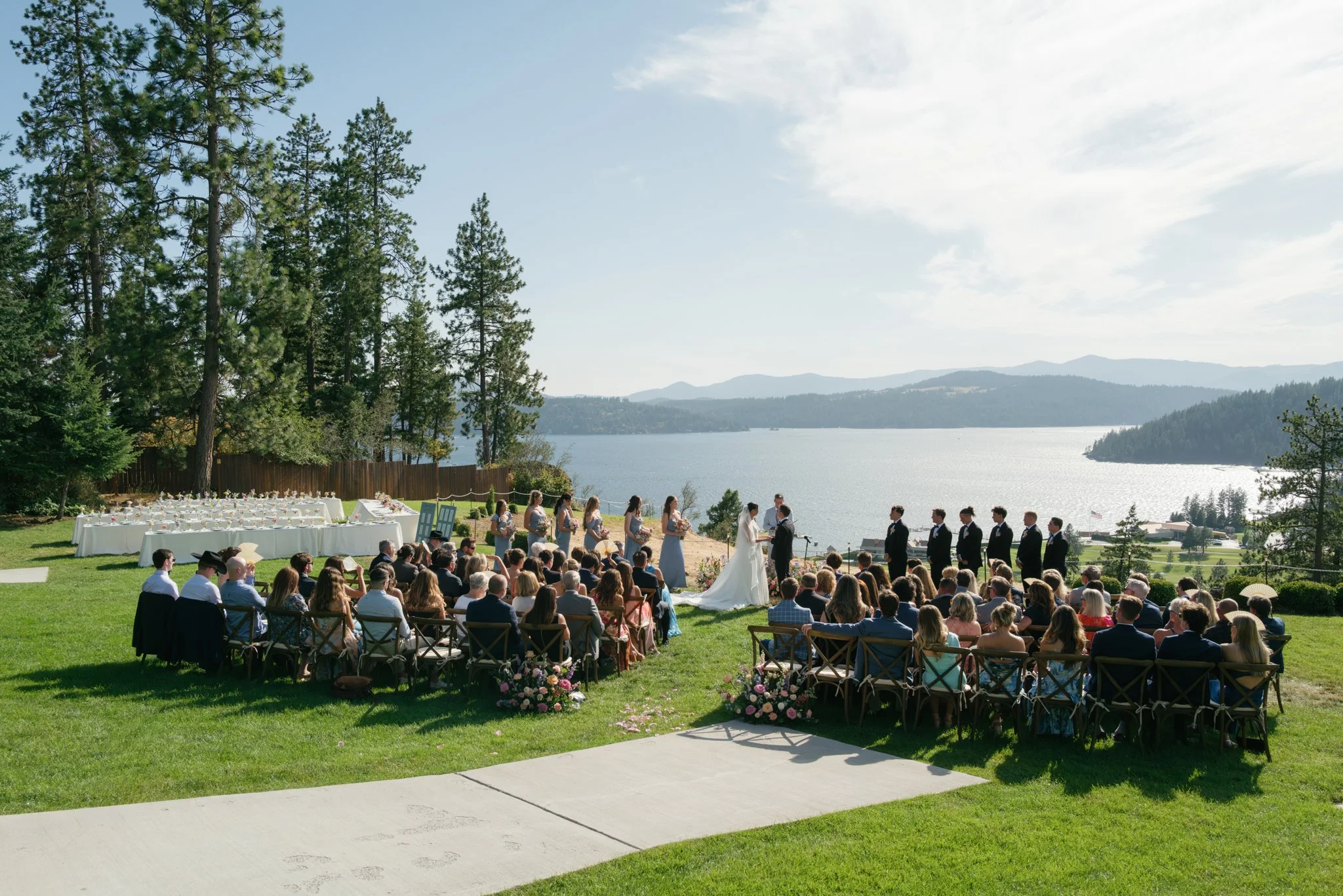 Outdoor wedding ceremony by a lake, with guests seated on chairs, a bride and groom exchanging vows, and bridesmaids and groomsmen standing nearby, surrounded by trees and scenic mountains in North Idaho.