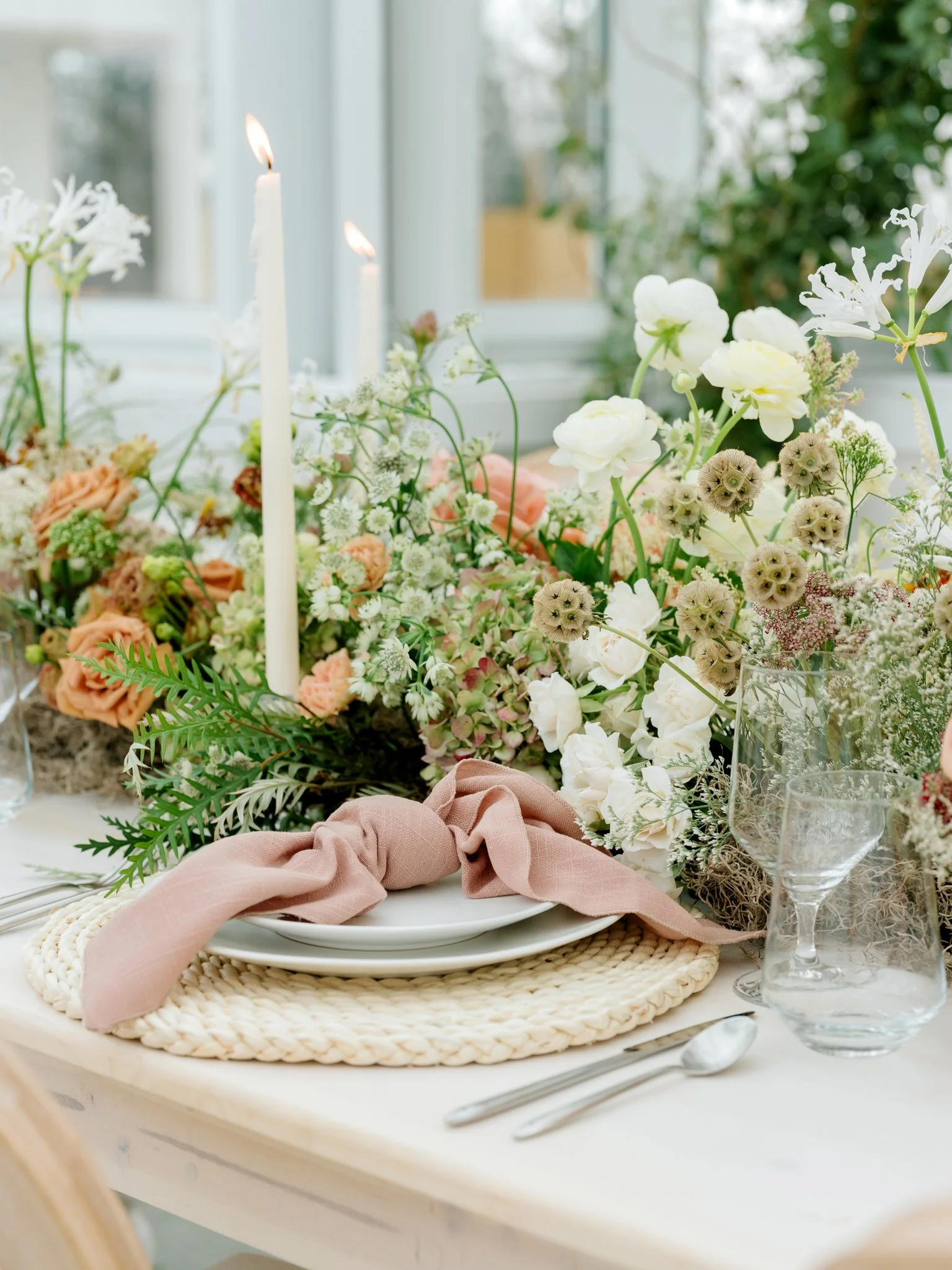 A table setting with white plates, a pink napkin, glassware, and surrounded by a floral centerpiece with white and peach flowers, two lit candles, and greenery.