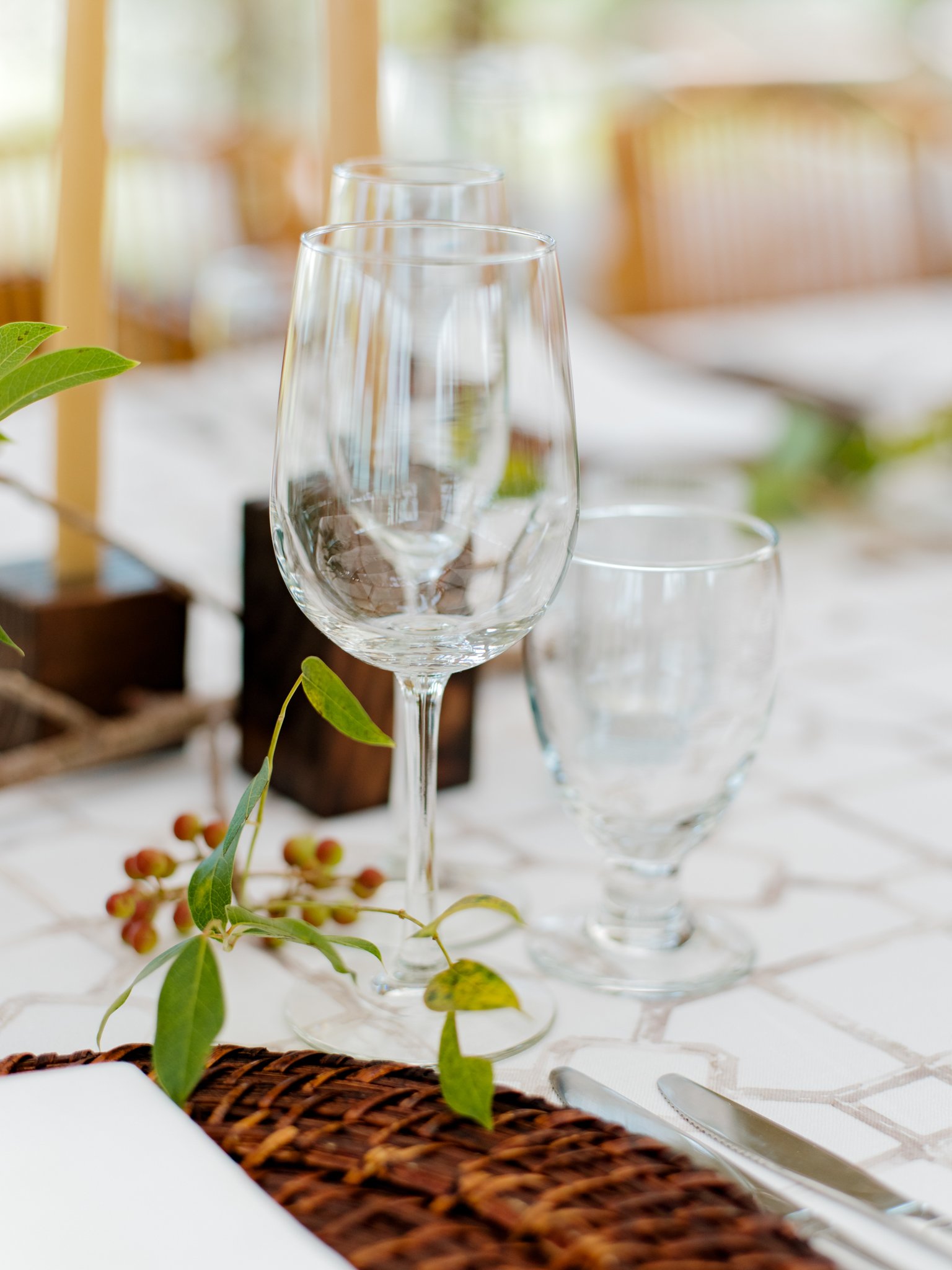 Empty wine glasses and a water glass on a dining table with a white tablecloth, a wicker placemat, and some greenery and berries in the foreground.