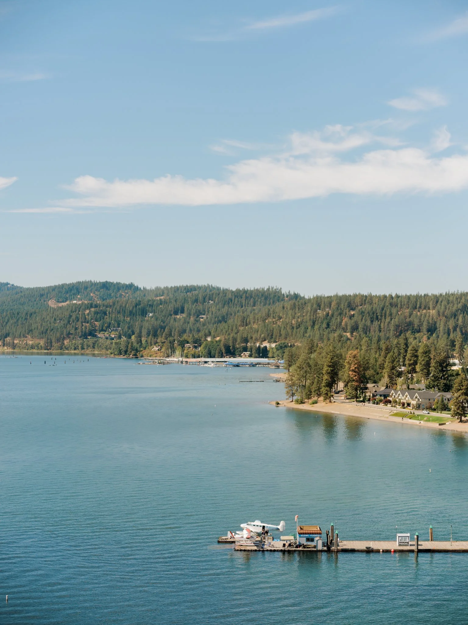 Wide aerial landscape of Lake Coeur d’Alene with a dock and surrounding forested shoreline on a clear summer day.