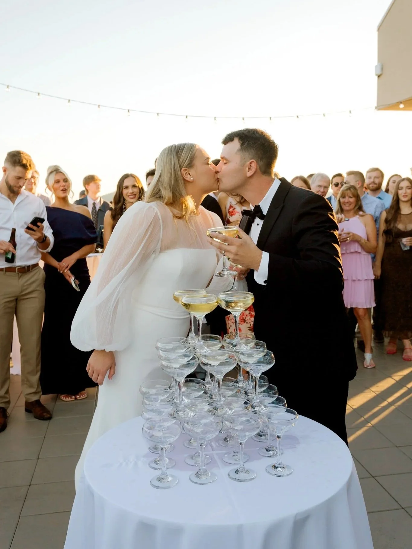 Bride and groom kissing during a wedding reception with guests in the background, standing around a table with a pyramid of champagne glasses at the Hagadone Event Center in Coeur d'Alene, Idaho.
