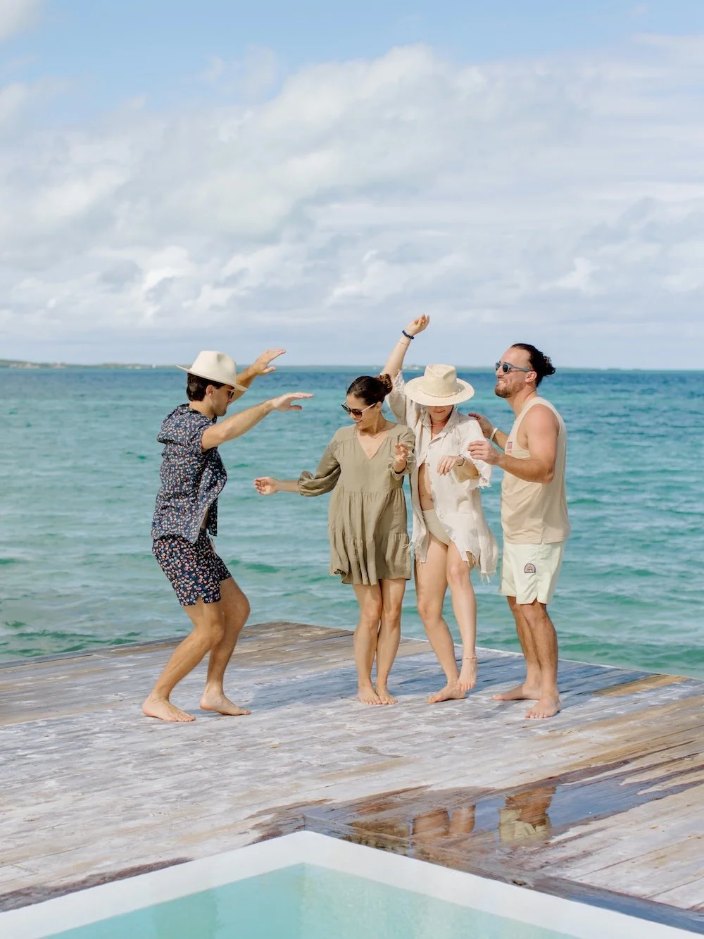 Four friends dancing on a wooden dock by the ocean on a cloudy day, dressed in casual summer clothes and hats.