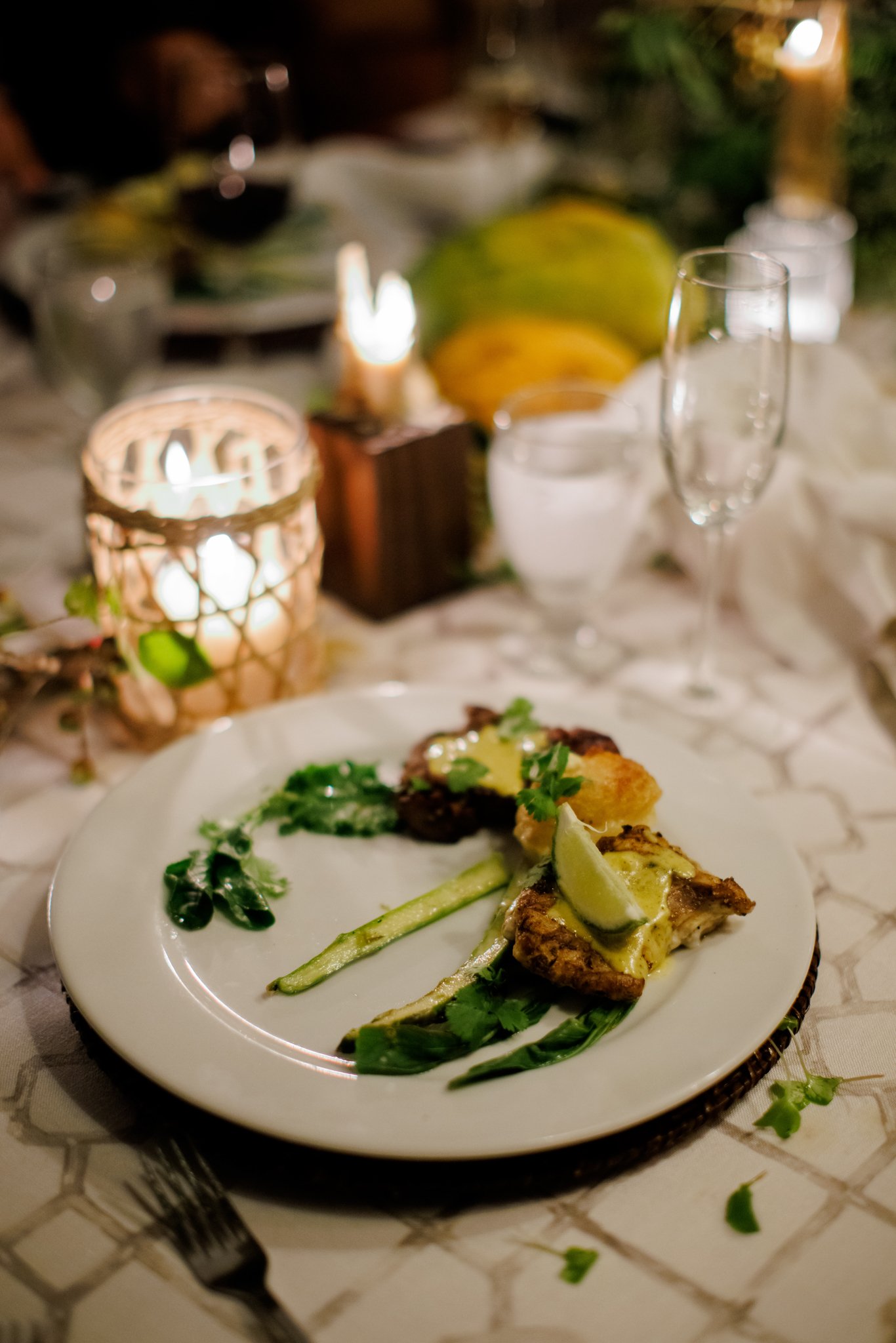 A dinner plate with grilled chicken, vegetables, and garnishes, on a decorated table with candles and glasses.