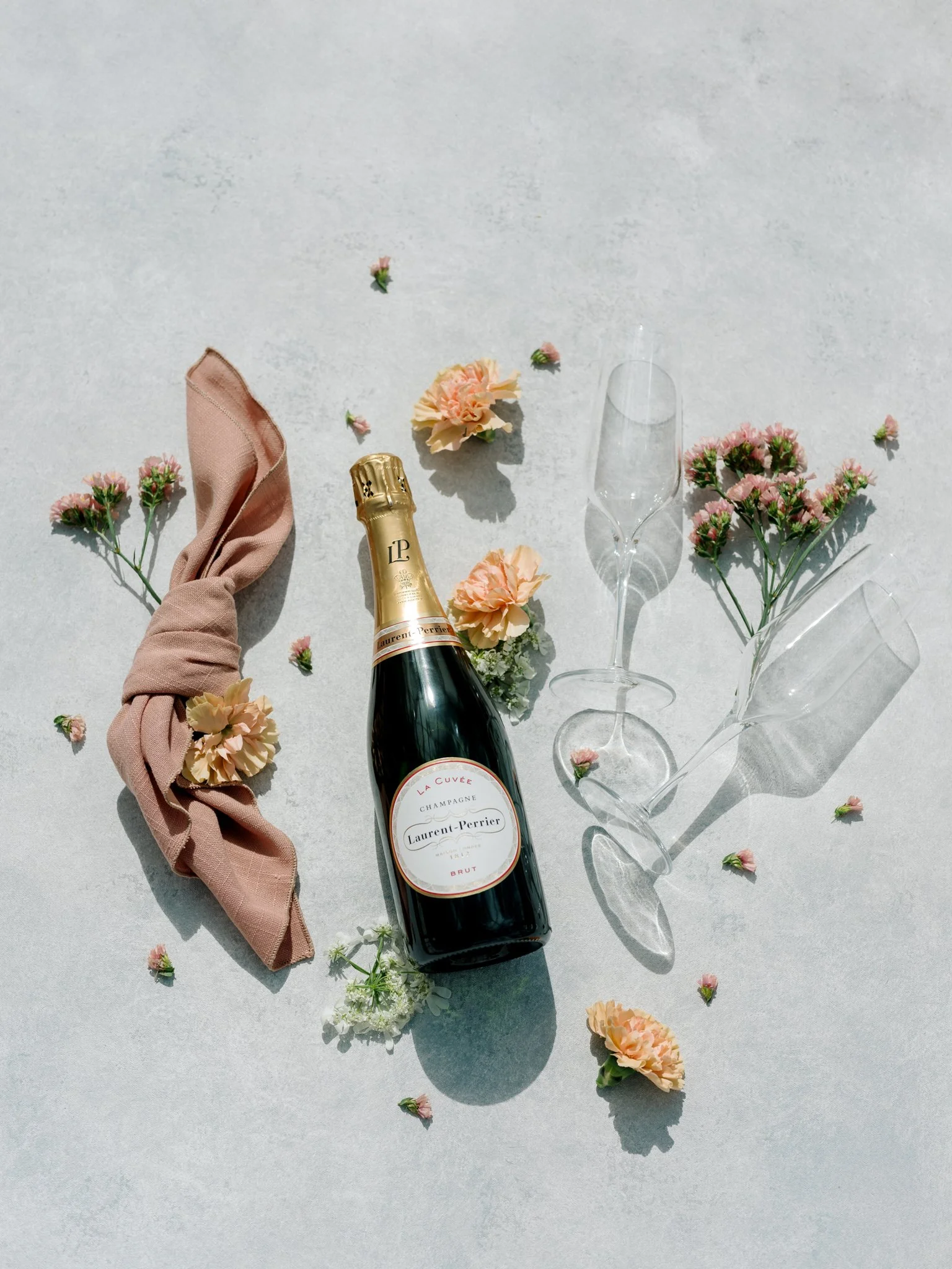 A bottle of Laurent-Perrier champagne, two empty champagne flutes, pink carnations, a pink cloth napkin, and pink flower petals on a light-colored surface at Greenhouse on Harvard near Spokane, Washington.