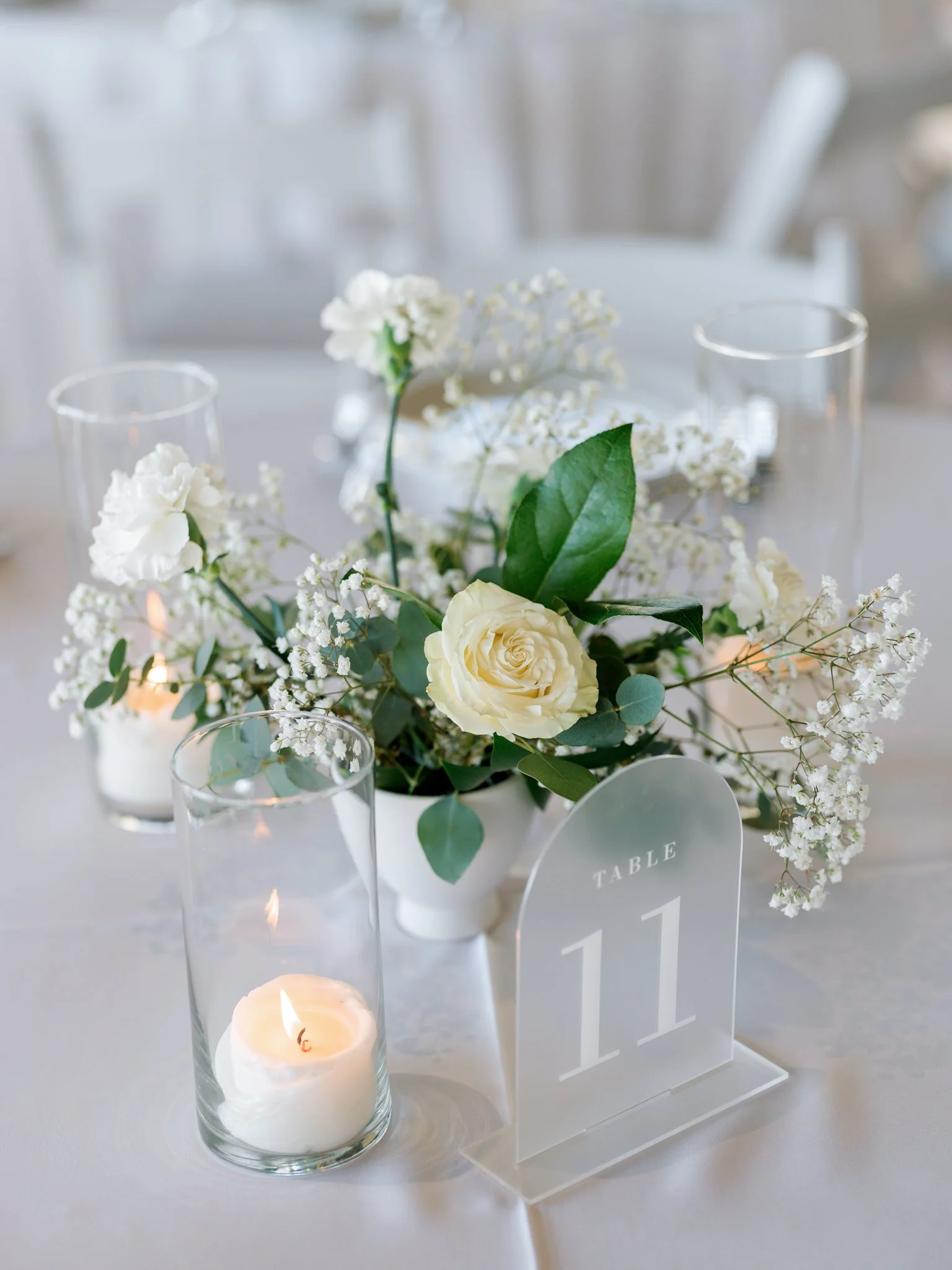 A wedding table centerpiece with white roses, baby's breath, green leaves, a table number card labeled 11, and candles in glass holders on a white tablecloth.
