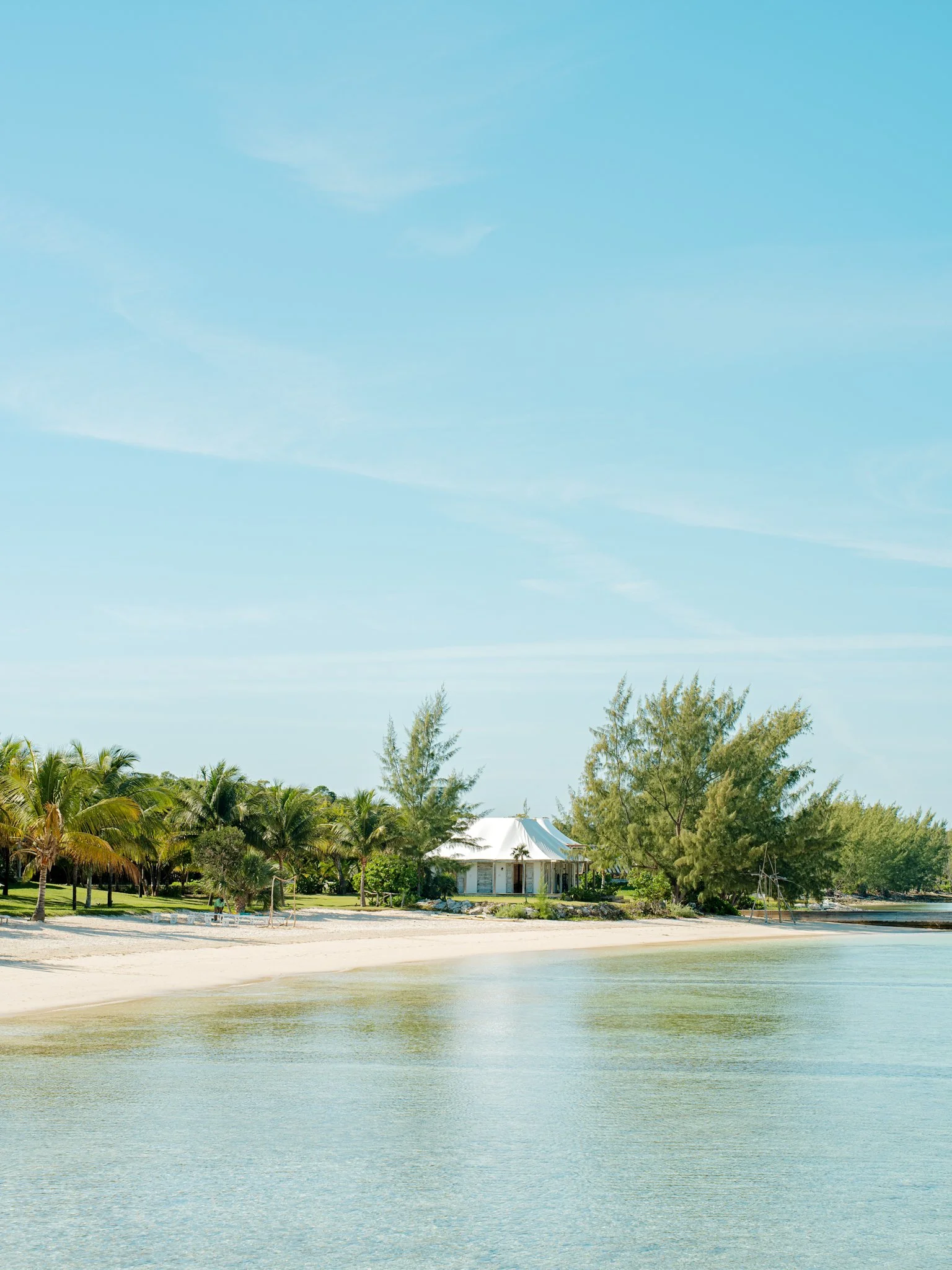 A tropical beach with calm water, sandy shore, palm trees, and a white building with a tented roof in the background under a clear blue sky.