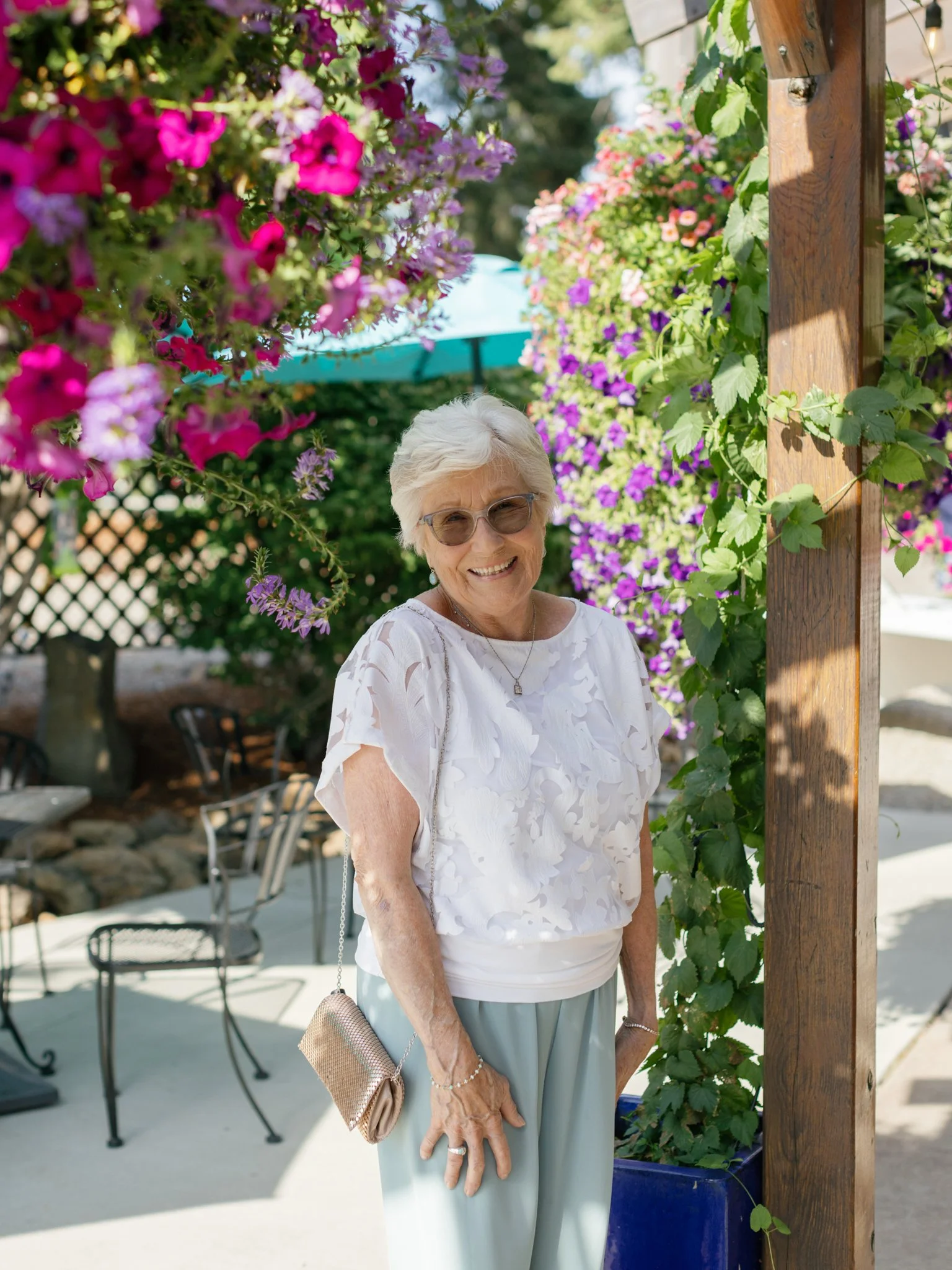 Smiling elderly woman with white hair, sunglasses, and jewelry, standing outdoors surrounded by colorful flowers and greenery.