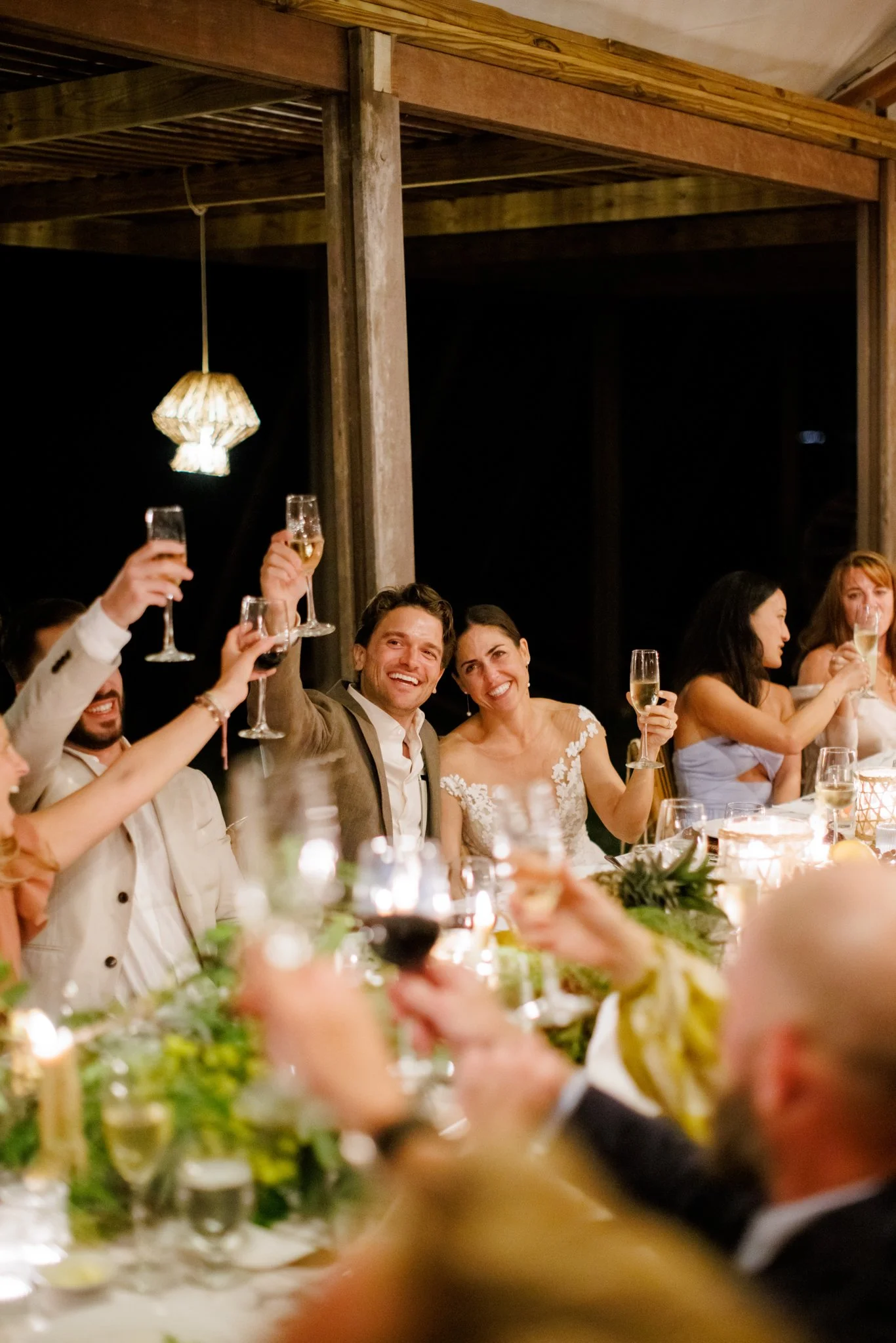 People at a wedding celebration raising glasses for a toast, smiling, seated at a decorated dining table with candles and greenery.