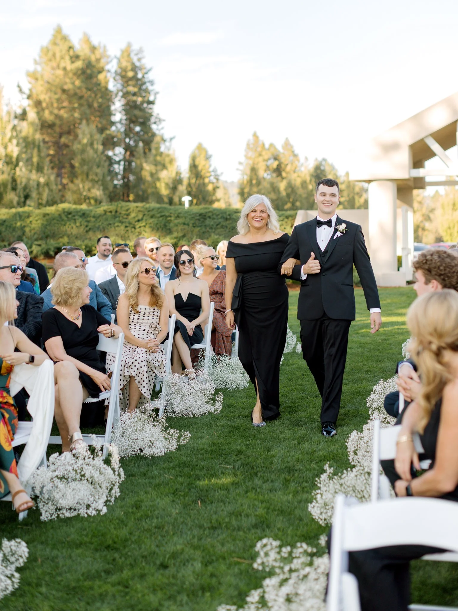 Groom escorting his mother down the aisle past seated guests during an outdoor ceremony at the Hagadone Event Center.