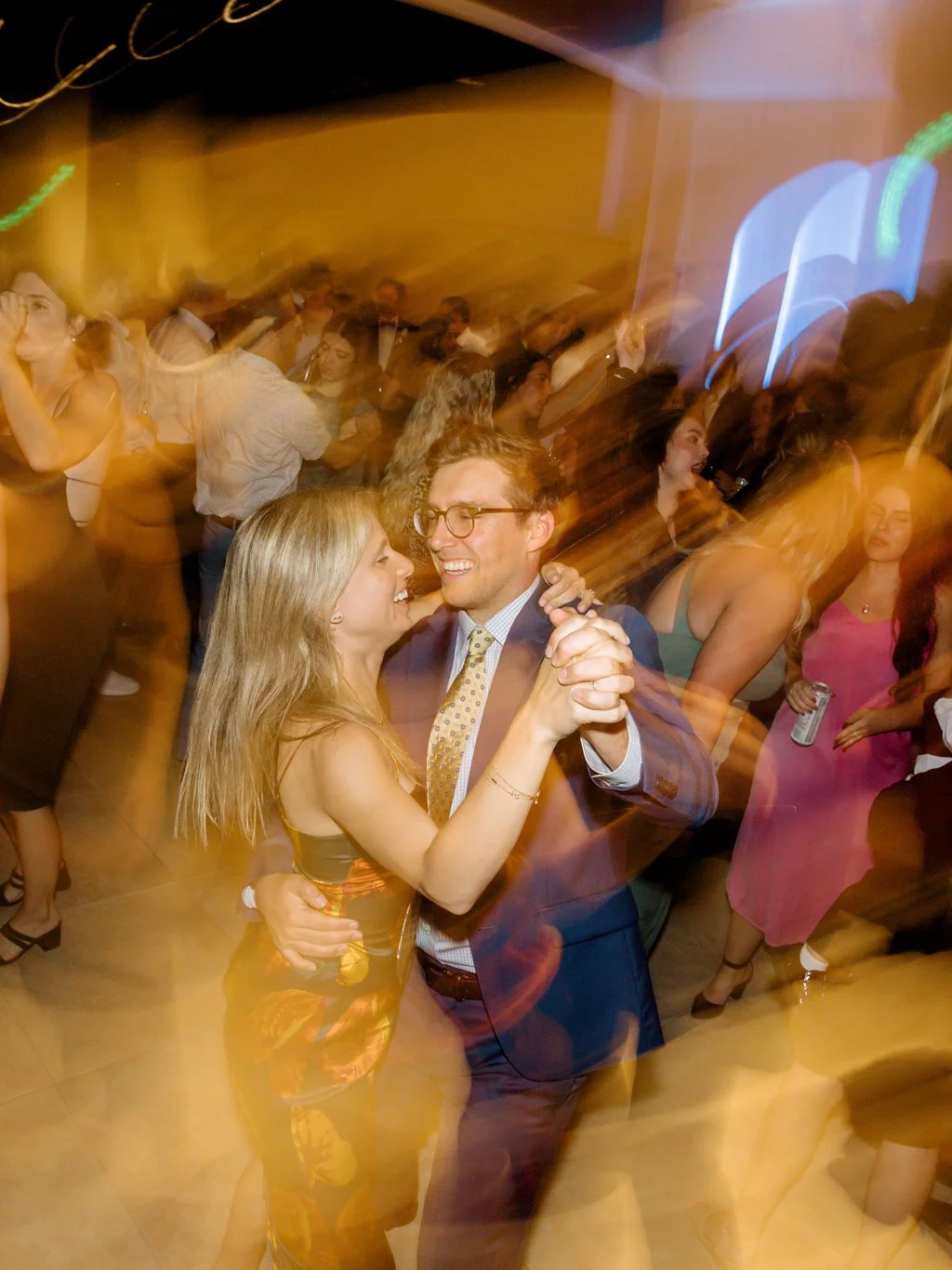Couple dancing together and smiling at a lively party with many people dancing around, colorful lighting, and a festive atmosphere.