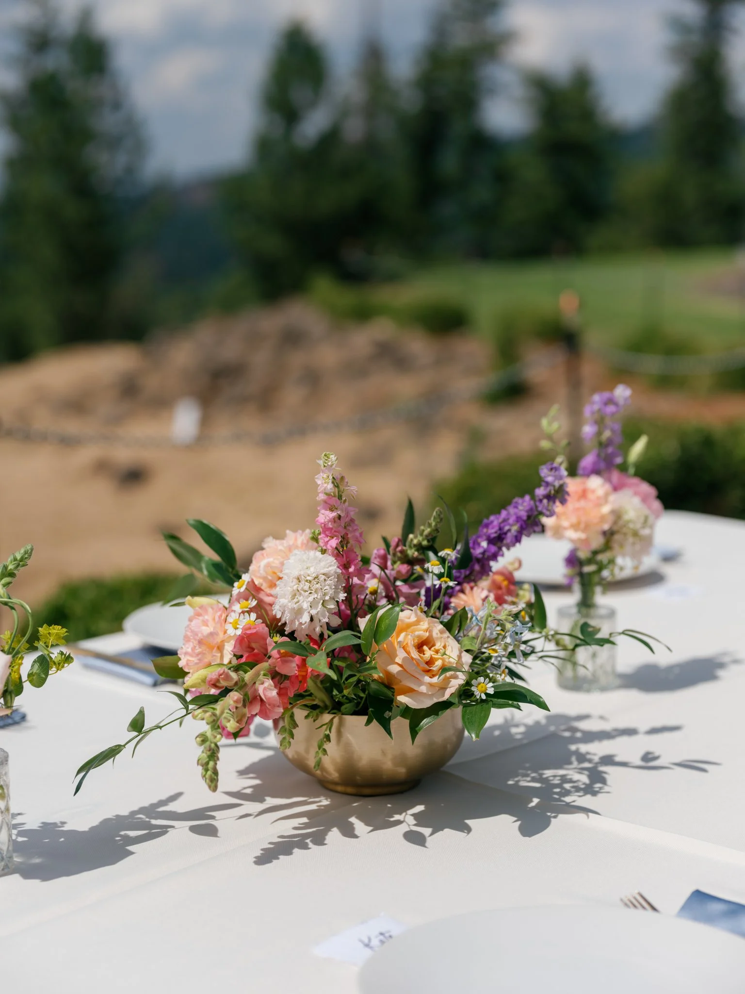 A table setup outdoors with a floral centerpiece in a metallic bowl, featuring pink, peach, white, and purple flowers, with a blurred natural background at Elk Point wedding above Lake Coeur d’Alene.