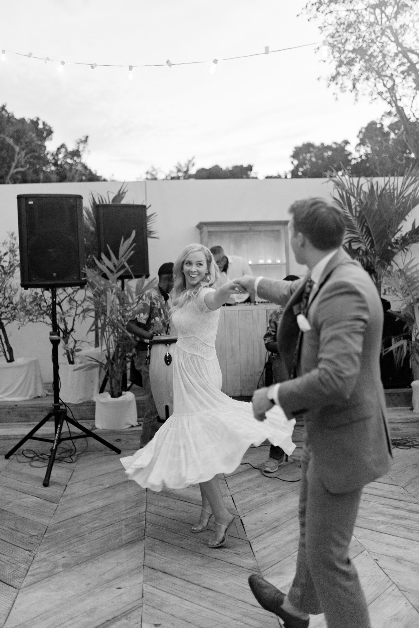 A woman in a white dress and a man in a suit dancing at an outdoor party with string lights above, palm plants in the background, and a DJ setup behind them.