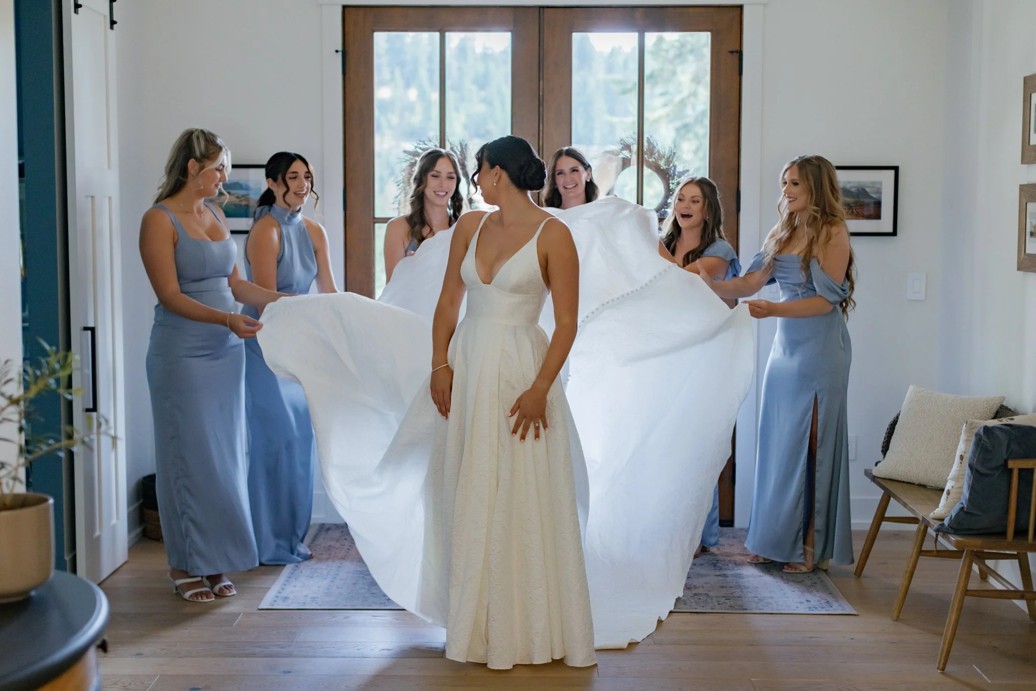 A bride in a white dress is surrounded by six bridesmaids in light blue dresses inside a room with wooden floors and large windows, as they prepare for her wedding.