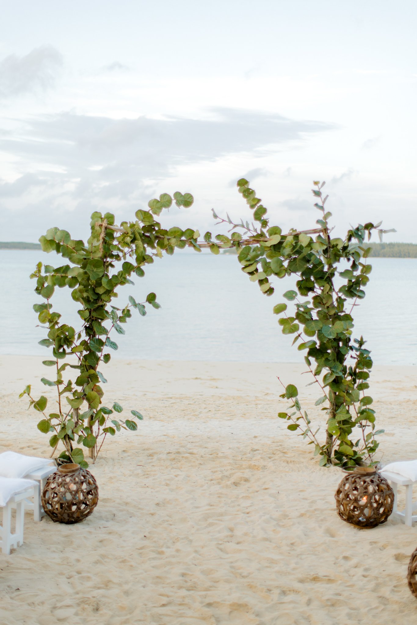 A decorative wedding arch made of green leafy branches on a sandy beach with a body of water in the background at The Other Side Hotel Eleuthera during a luxury Bahamas destination wedding.