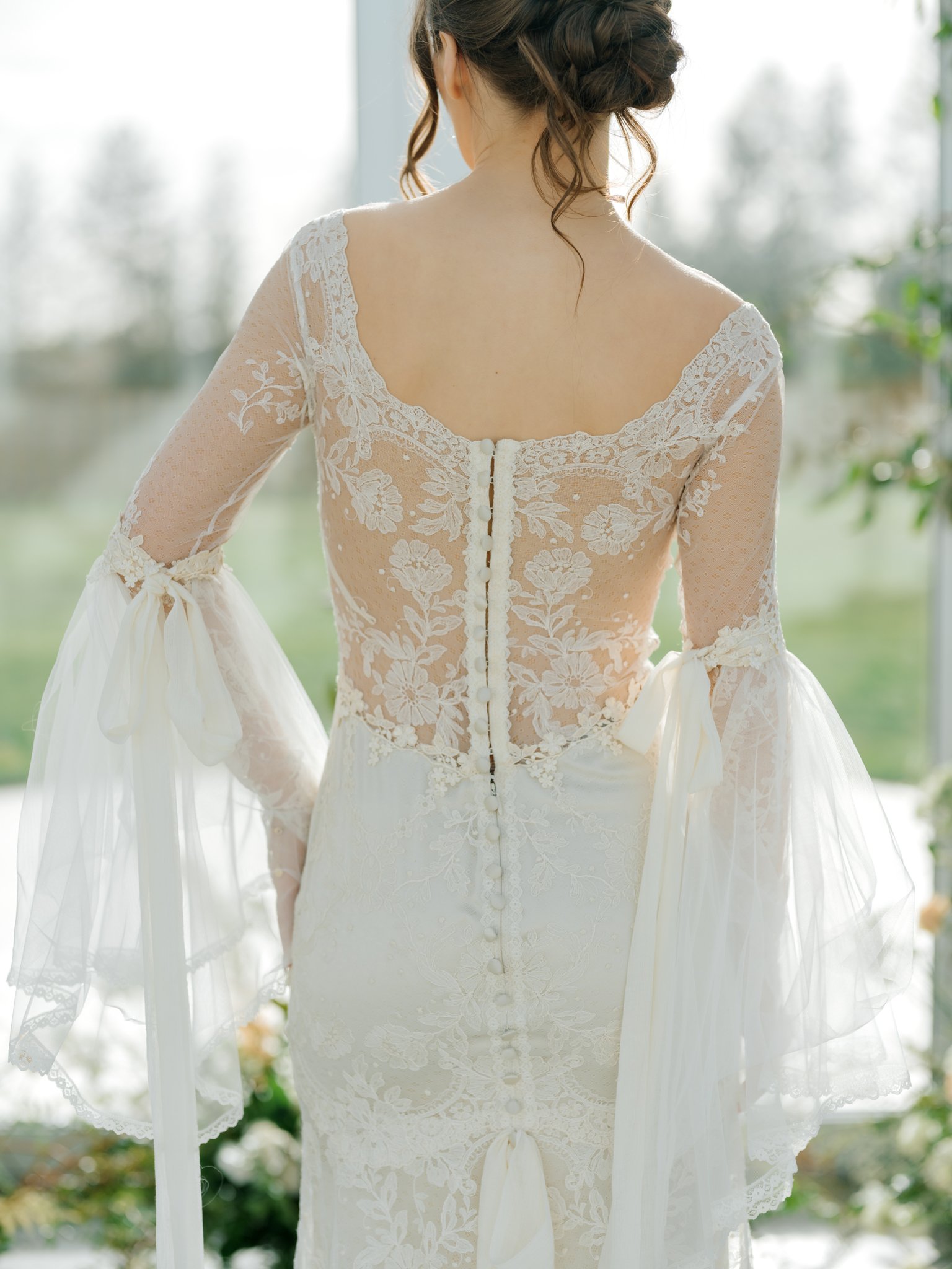 Back view of a bride in a white lace wedding dress with floral patterns, long lace sleeves with ribbon ties, and a row of fabric-covered buttons down the back, standing by a window with a blurred outdoor background.