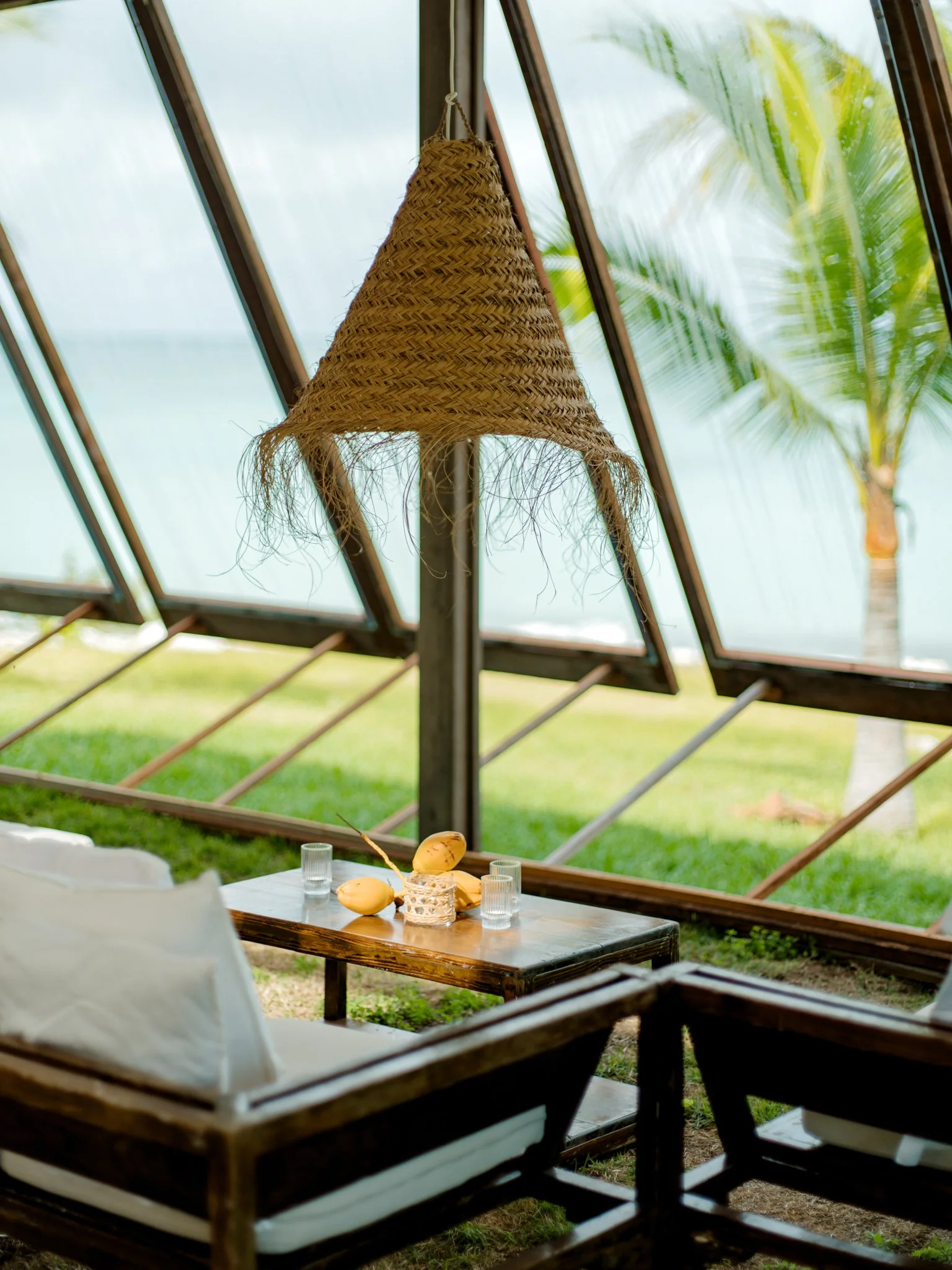 Outdoor seating area with a wooden table, glass cups, and decorative yellow gourds, under a woven hanging lamp, with a view of a tropical palm tree outside via large slanted windows.