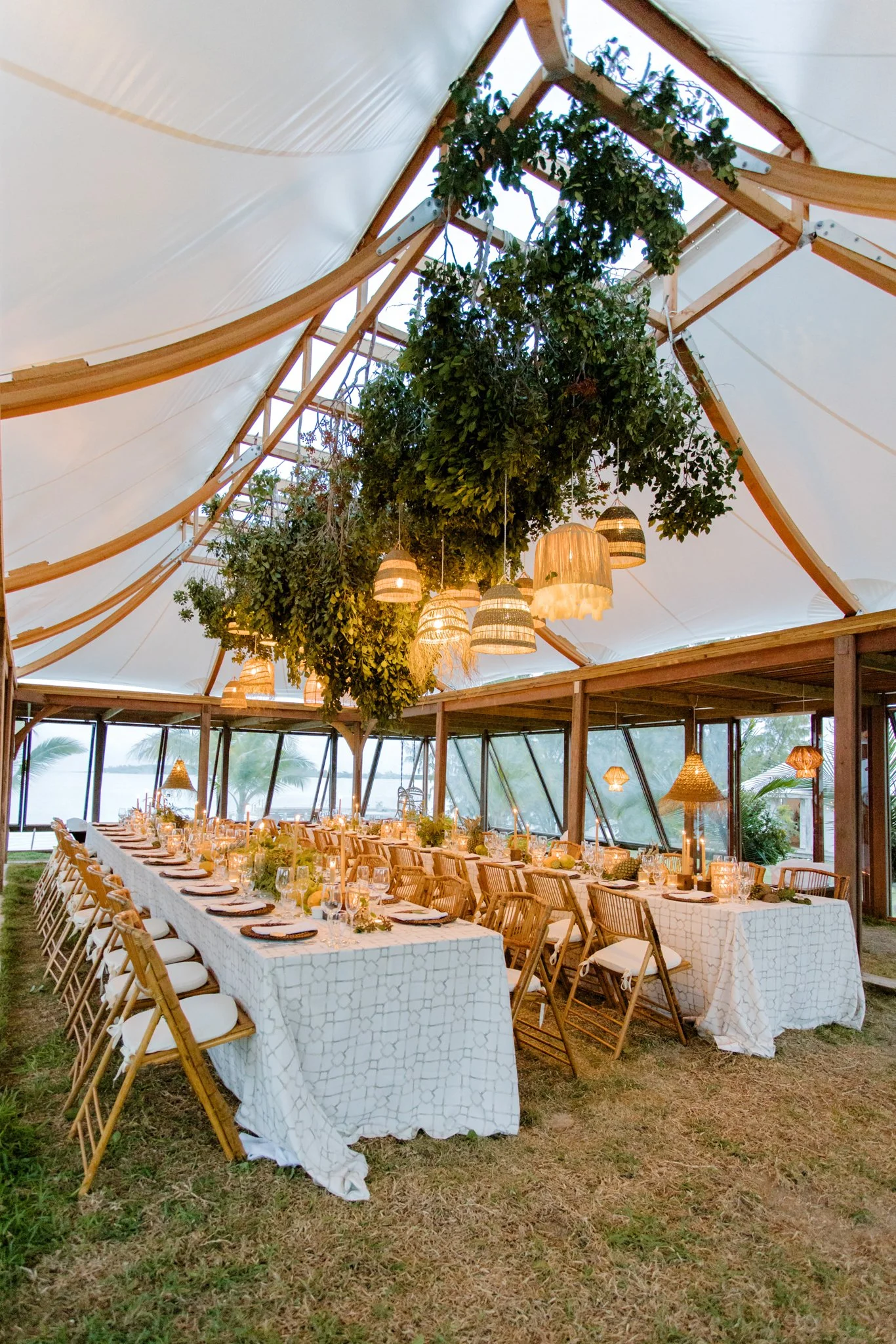 An elegant outdoor dining setup under a tent with long white tablecloths, wooden chairs with white cushions, and hanging woven lamps surrounded by lush greenery and tropical landscape near water.