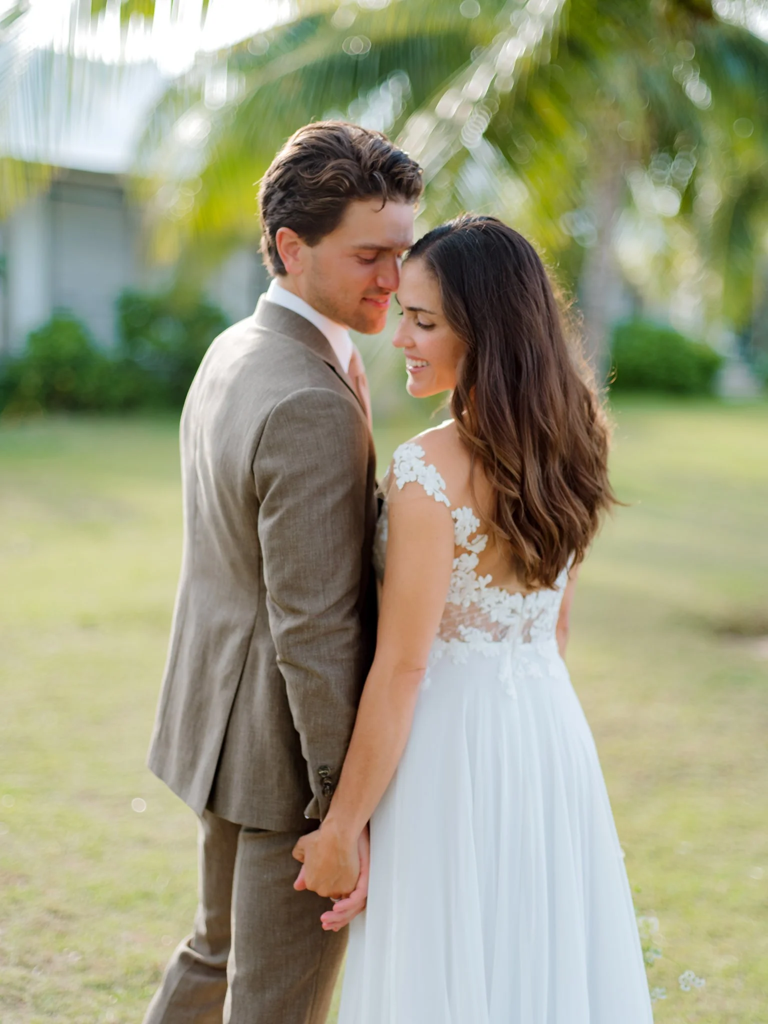 A bride and groom standing close outdoors, holding hands, with their foreheads touching and smiling, green blurred background, bride in a white gown with lace details, groom in a light brown suit at The Other Side Hotel Eleuthera Wedding.