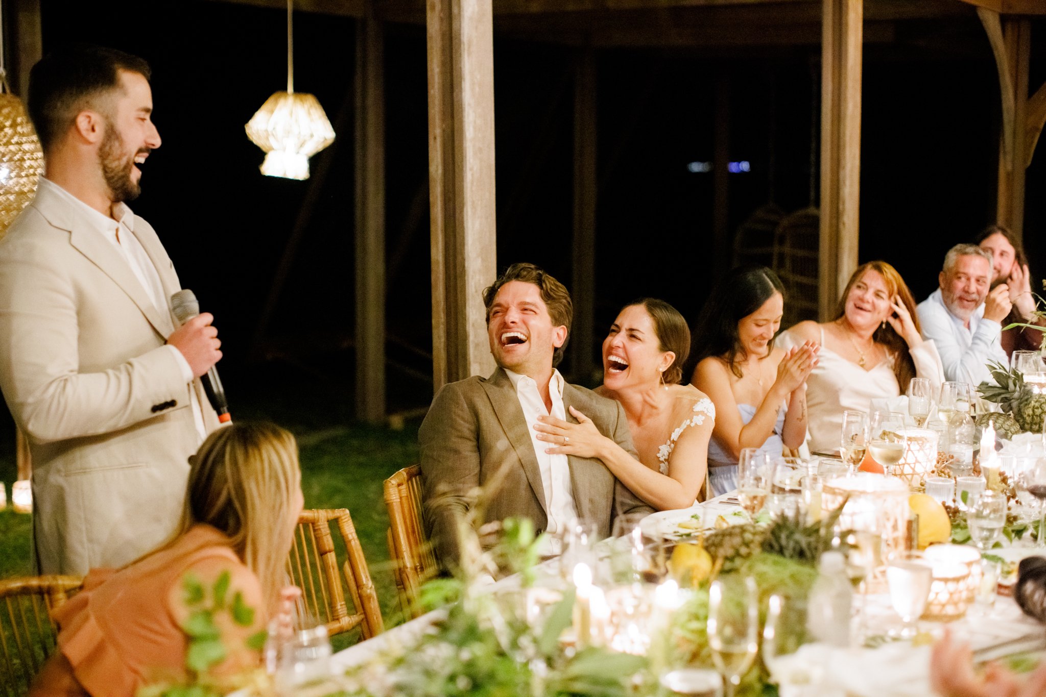 A man in a cream-colored suit is giving a speech at a wedding reception, holding a microphone and smiling. Guests are seated at a long table, laughing and enjoying the moment, with some clapping and one with hand on chest, in a decorated outdoor venue at night.