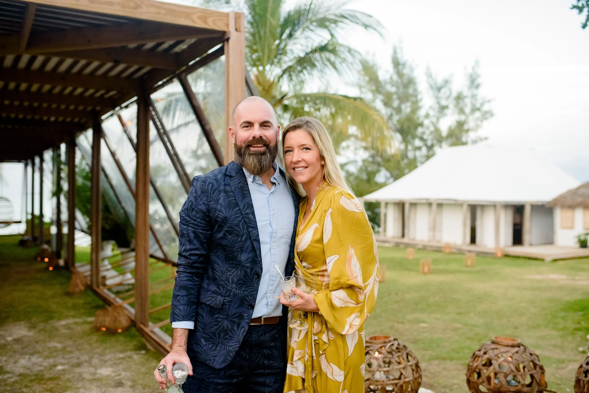 A smiling man with a beard and a woman with blonde hair stand close together outdoors during daytime, holding drinks. Behind them, there are palm trees, a wooden structure, and a building under construction, suggesting a tropical or beachside setting.