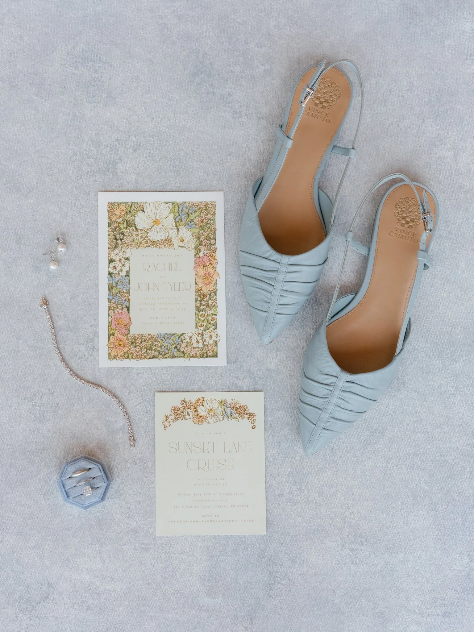 A pair of light blue high-heeled shoes, wedding invitation cards, a pearl necklace, and a ring in a blue jewelry box are arranged on a light gray surface.