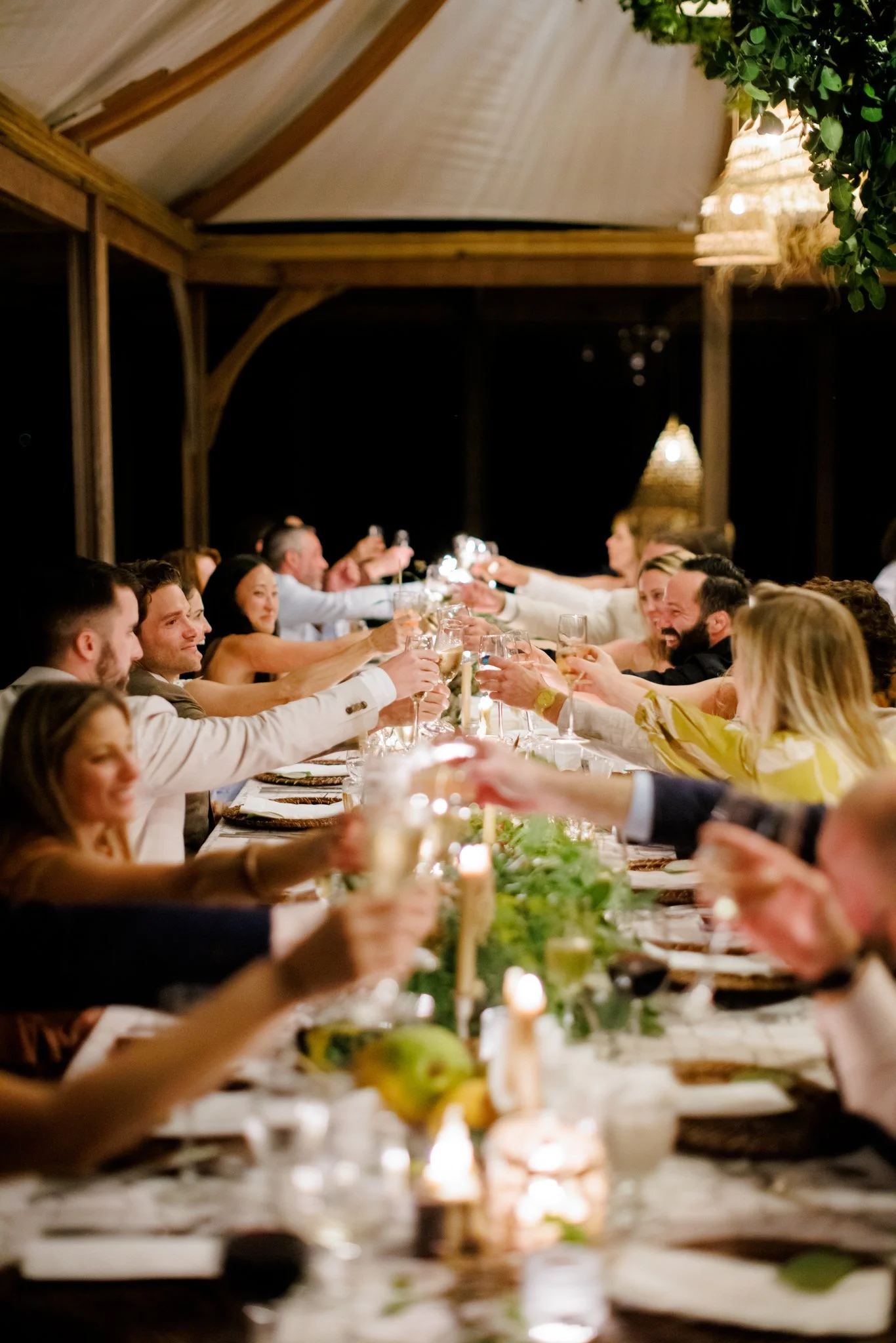 A group of people sitting at a long dinner table raising glasses for a toast during a celebration at night, under a wooden gazebo with hanging lights and greenery.