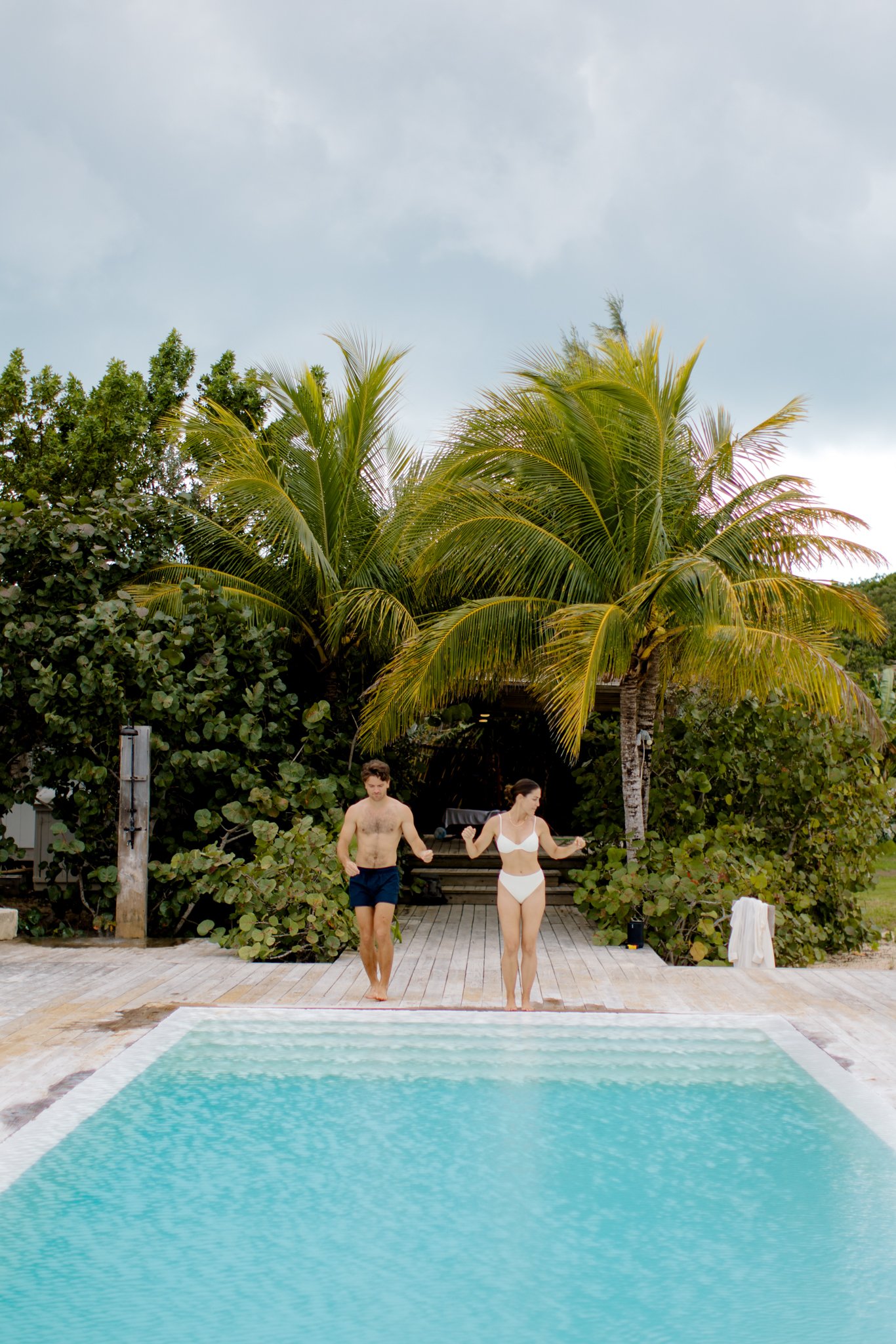 A man and woman in swimwear walking out of a swimming pool towards a tropical backdrop of palm trees and lush greenery at The Other Side Hotel during their destination wedding in Eleuthera, Bahamas.