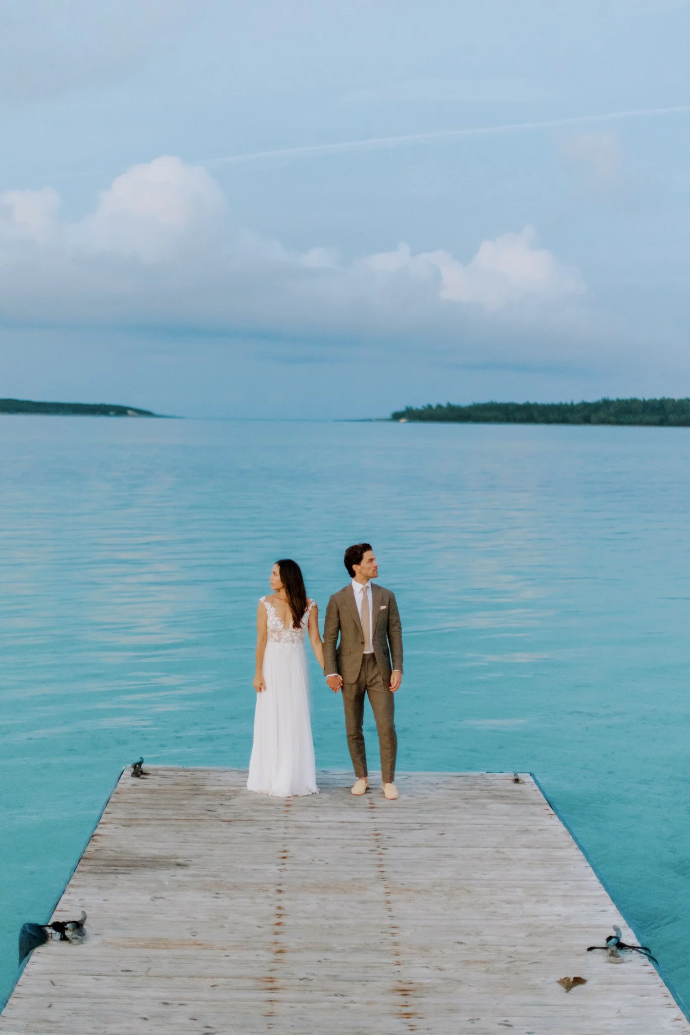 A bride and groom standing hand in hand on a wooden dock over calm turquoise water, with a cloudy sky in the background at The Other Side Hotel Eleuthera during a luxury Bahamas destination wedding.