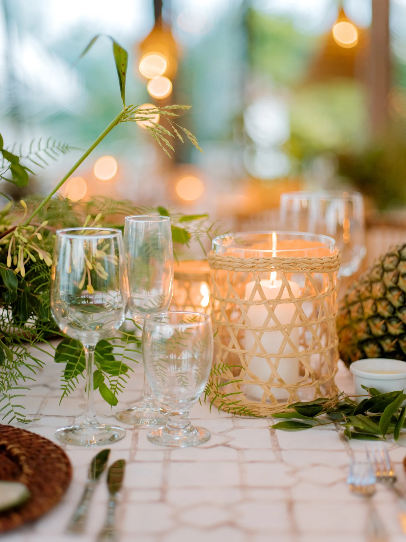 A table decorated with greenery, a candle in a woven holder, pineapple, and wine glasses at an outdoor setting with blurred string lights in the background.