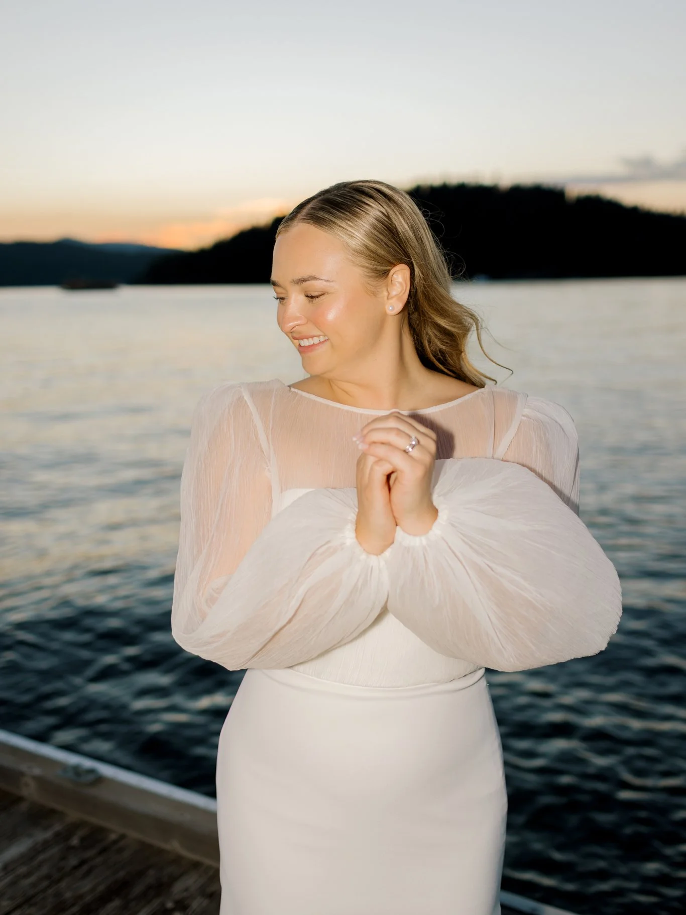 A smiling woman in a white dress and sheer long sleeves stands on a dock by the water at sunset, with her eyes closed and hands clasped in front of her at the Hagadone Event Center in Coeur d'Alene, Idaho.