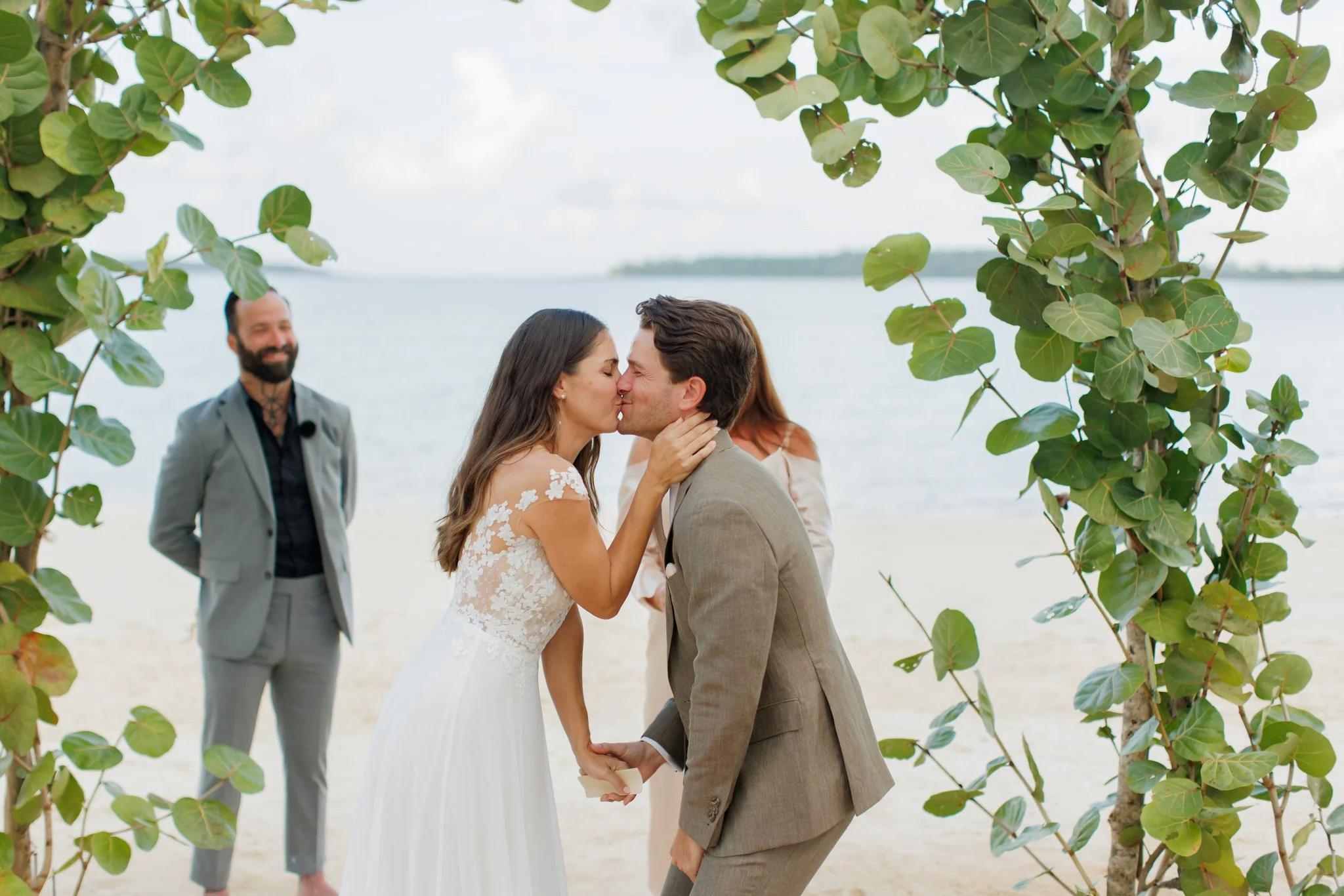 A couple is kissing during a beach wedding ceremony, with the bride in a lace wedding dress and the groom in a brown suit, held under leafy trees near the water. An officiant stands behind them smiling.