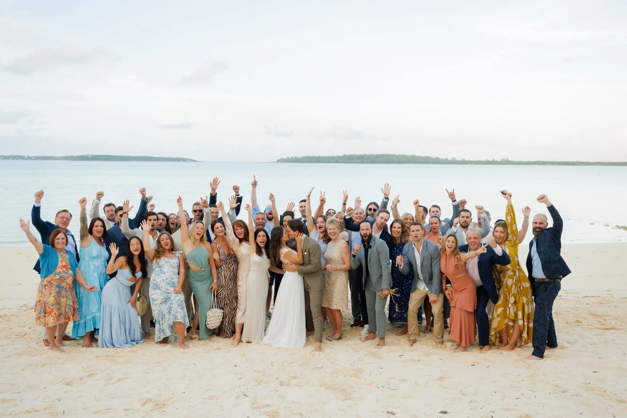 Group of wedding guests on a beach celebrating, with some raising their hands and making peace signs, in front of the ocean and cloudy sky.