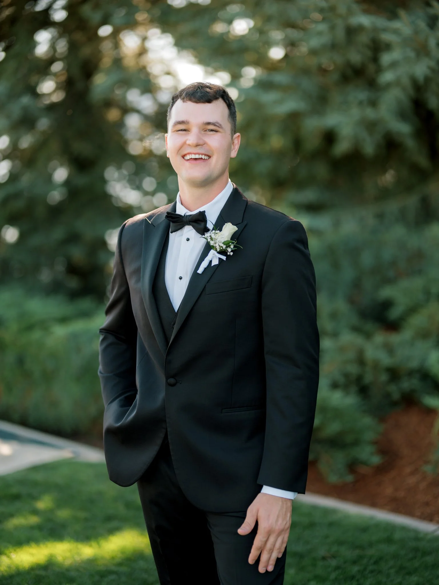 Groom smiling during solo portraits in a black tuxedo surrounded by evergreens at a Coeur d’Alene summer wedding.