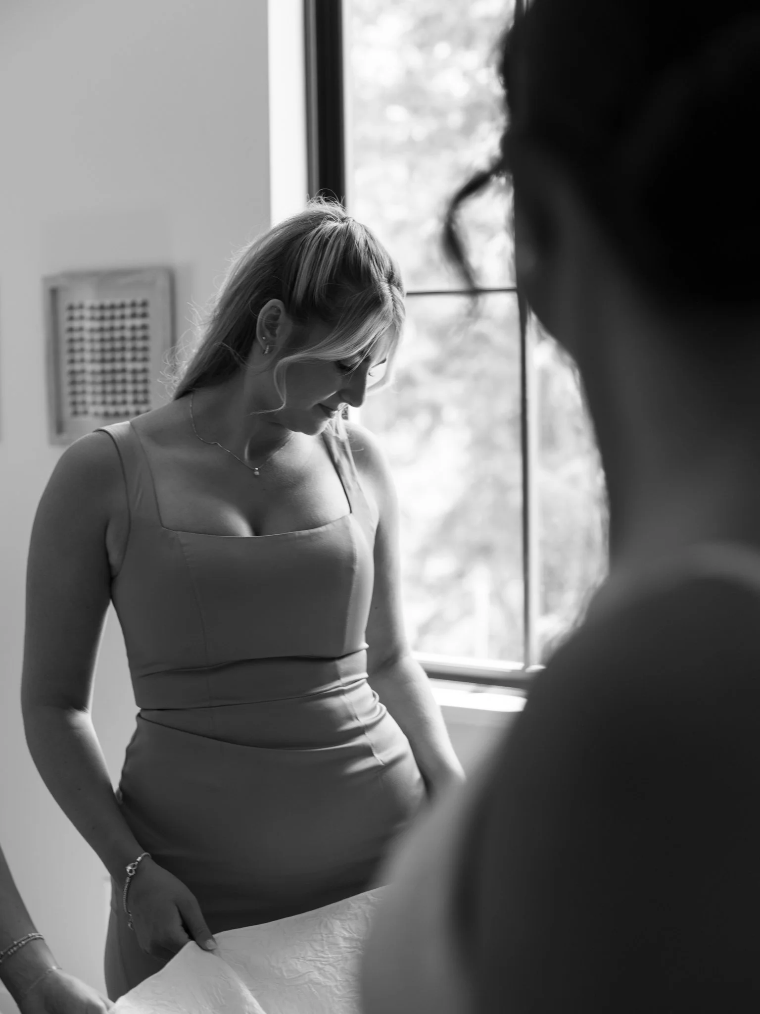 A black and white photo of a woman looking down, standing near a window with natural light illuminating her, and another person's shoulder and head in the foreground.