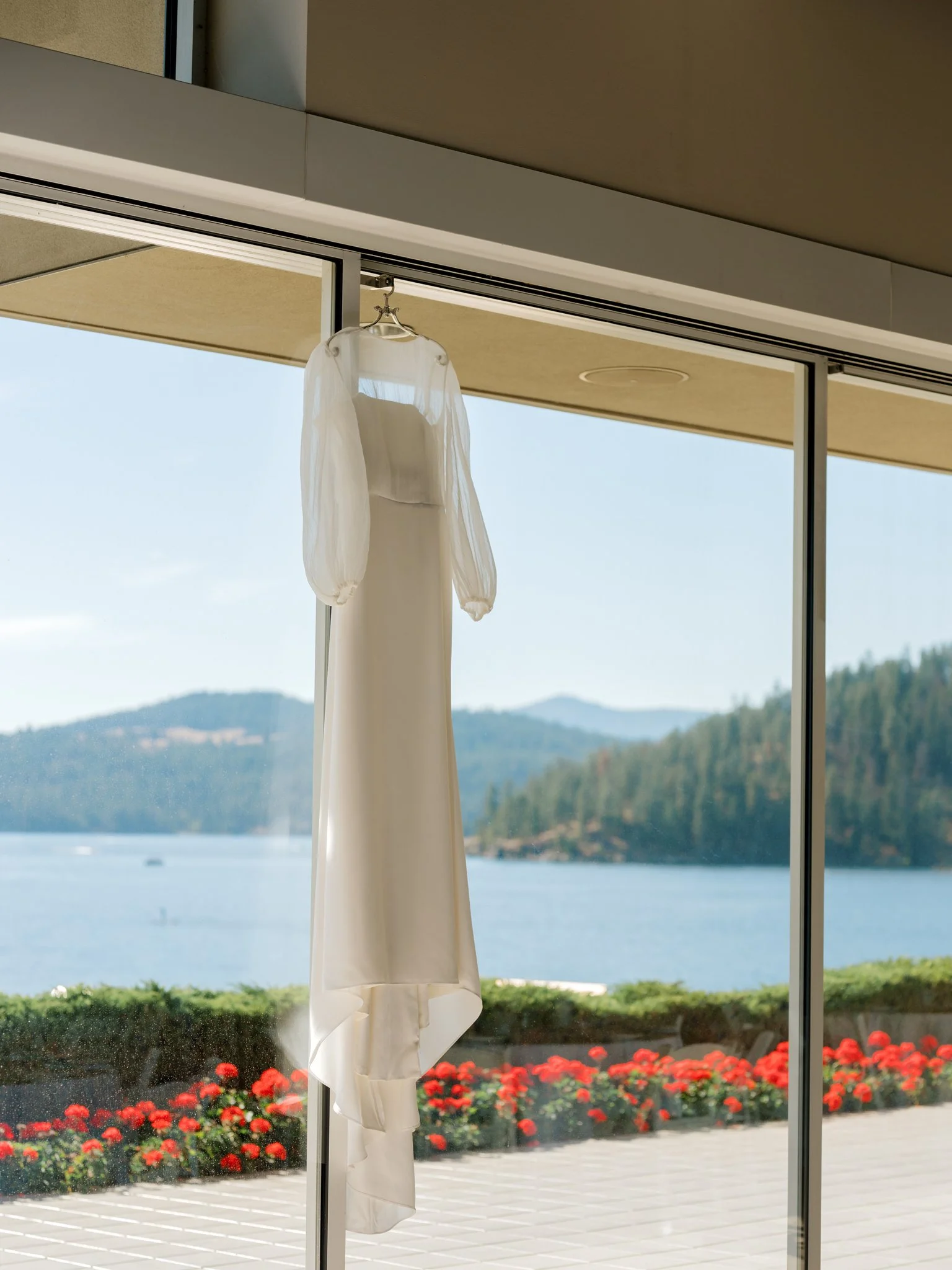 Bridal gown with sheer sleeves hanging in front of large windows overlooking Lake Coeur d’Alene at the Hagadone Event Center.