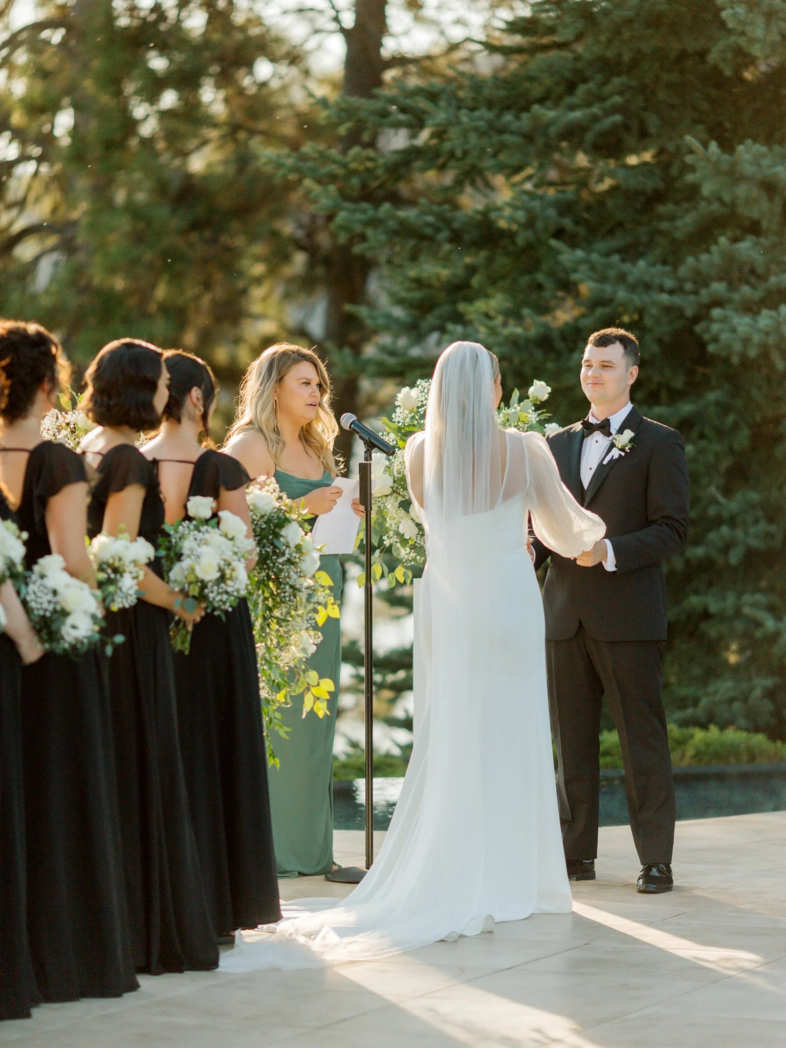 A wedding ceremony outdoors with a bride and groom holding hands, surrounded by bridesmaids and an officiant, with trees in the background at the Hagadone Event Center in Coeur d'Alene, Idaho.