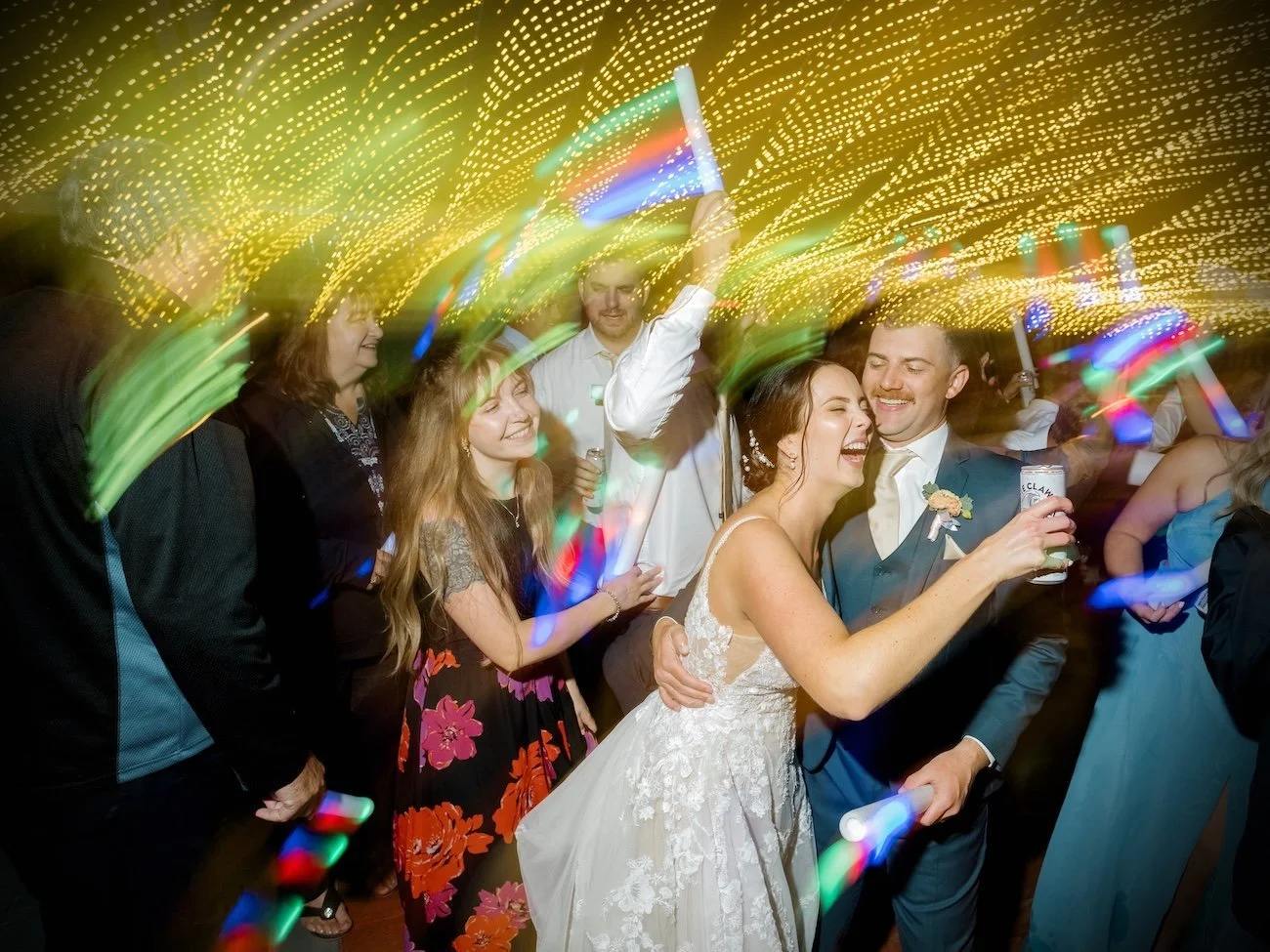 Happy bride and groom dancing with friends at a wedding reception, colorful lights blur overhead.