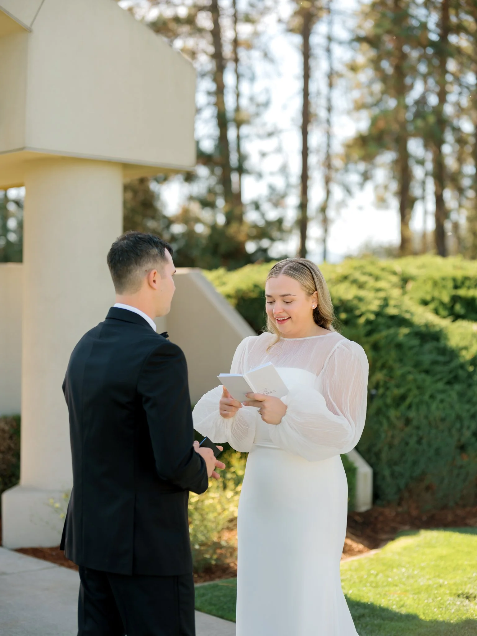 A woman in a white dress is reading vows to a man in a black suit during an outdoor wedding ceremony at the Hagadone Event Center in Coeur d'Alene, Idaho.